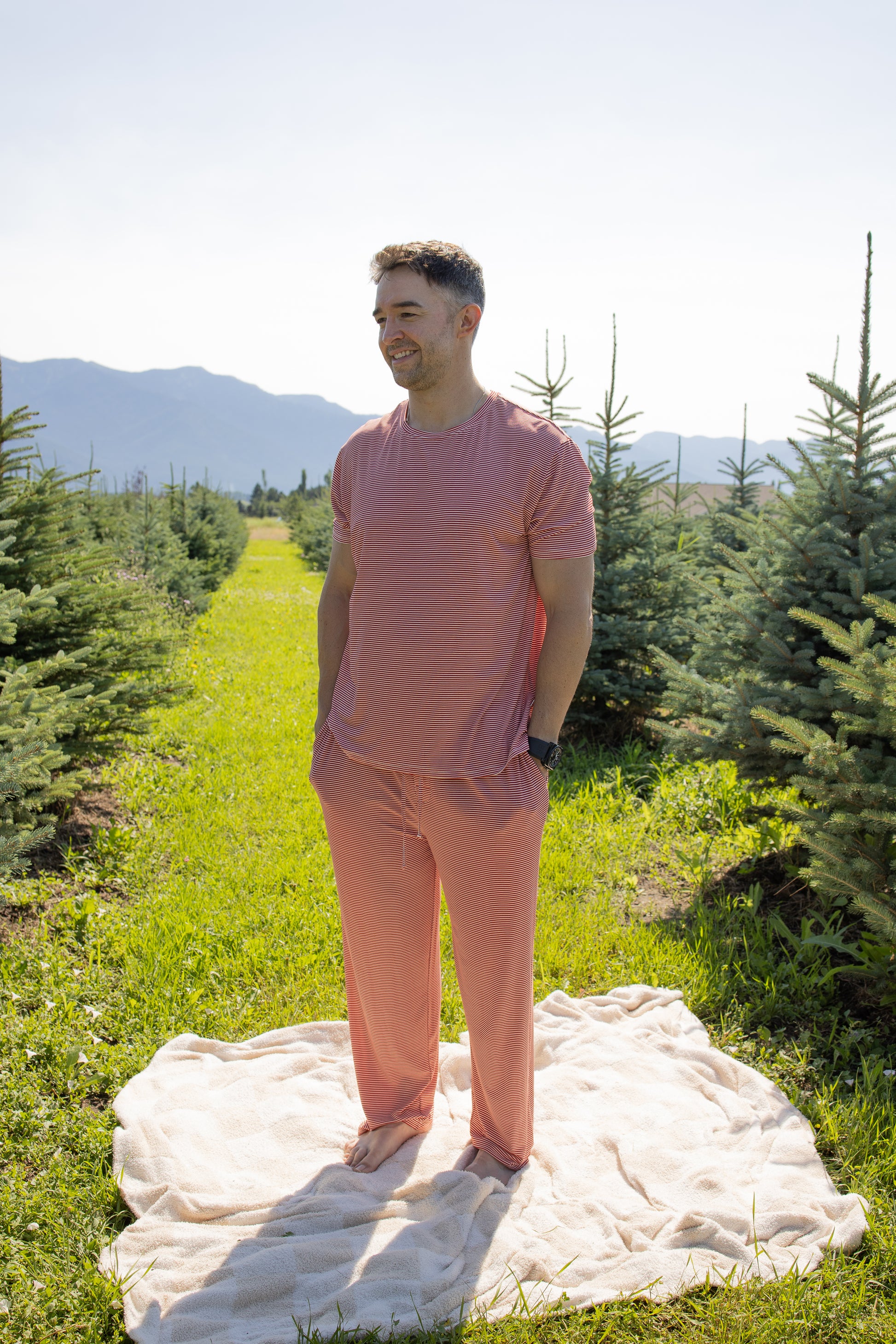 A man in the Men's Short Sleeve Pajamas in Peppermint Stripe Ribbed by forever french baby stands on a beige blanket between rows of pine trees in a sunny outdoor setting, with mountains in the background.