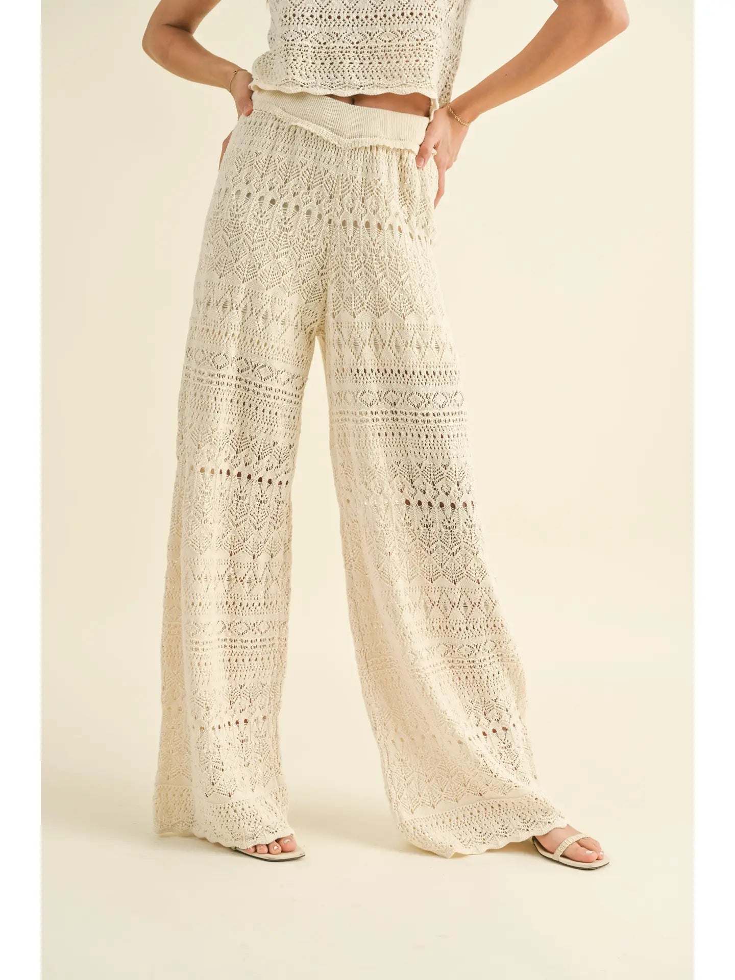 Dressed in the Crochet Pattern Pants from MIOU MUSE, a person wears a sleeveless crochet top crafted from soft cotton fabric accompanied by matching wide-leg pants in light cream. Standing against a plain backdrop with one arm bent and hand on hip, they pair their ensemble with open-toed sandals. To ensure these garments last, it's recommended to hand wash them.