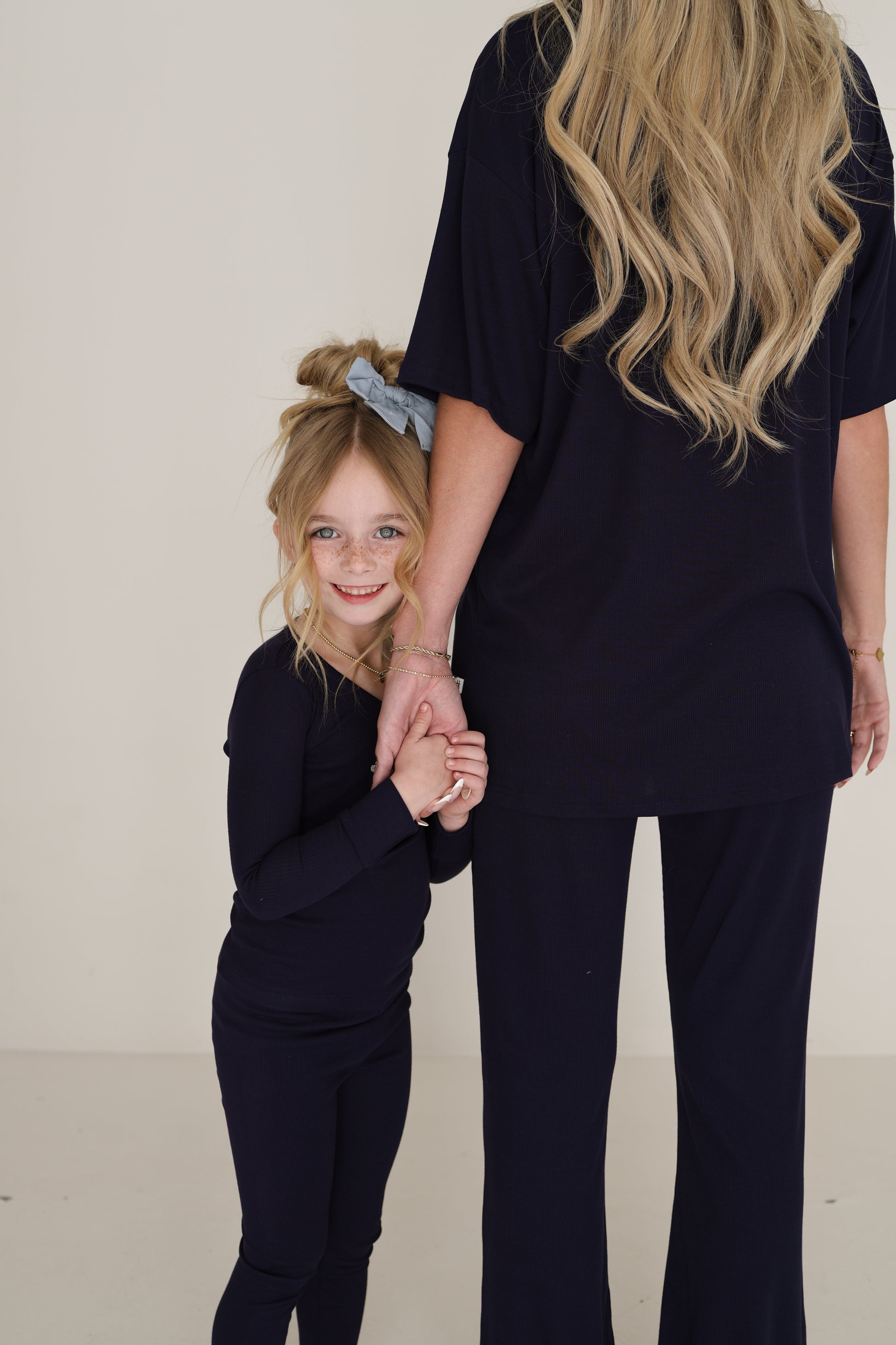 A young girl with long blonde hair and a blue bow smiles while holding hands with a woman, both wearing matching forever french baby Bamboo Two Piece Pajamas in Blueberry Ribbed. The woman is seen from the back.