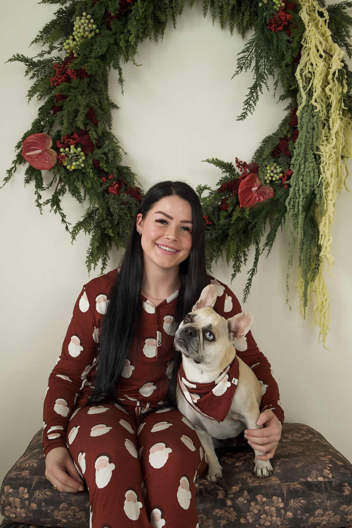 A woman in matching bamboo viscose pajamas sits with a French bulldog wearing a Bamboo Bandana | Vintage Santa in front of a holiday wreath.