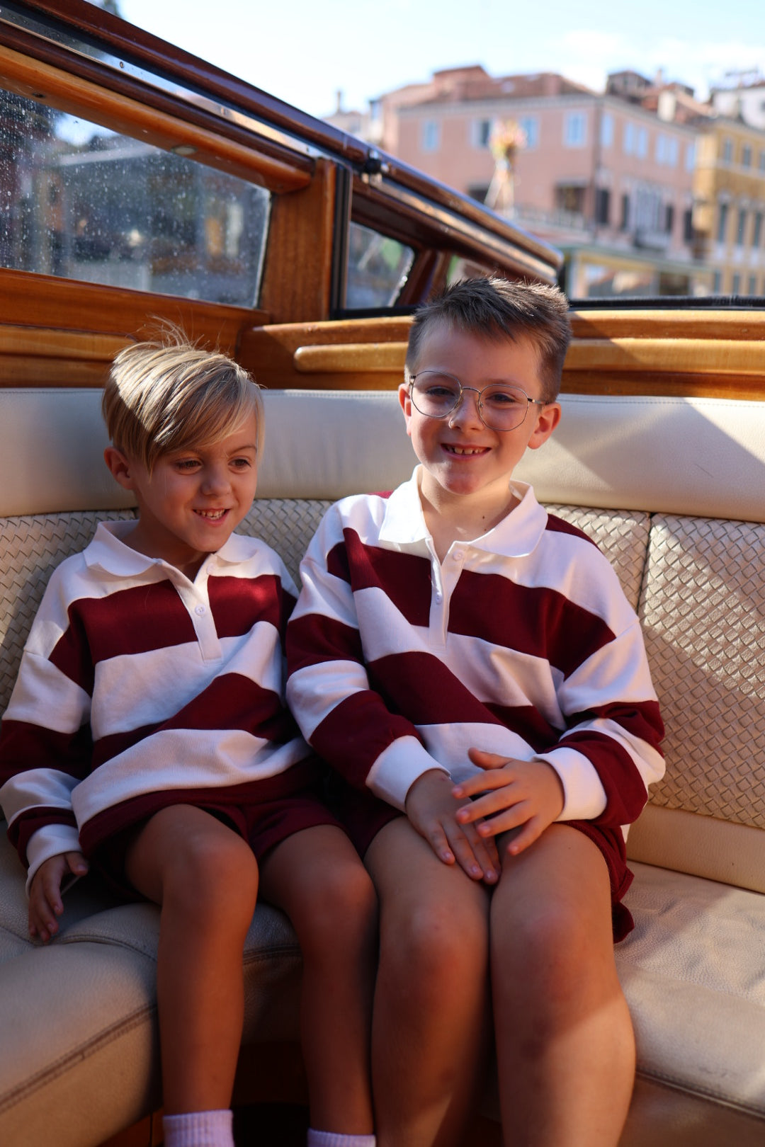 Two young boys sit side by side on a boat, both wearing matching Child Rugby Sweatshirt | Deep Cranberry tops with wide white and deep red horizontal stripes.