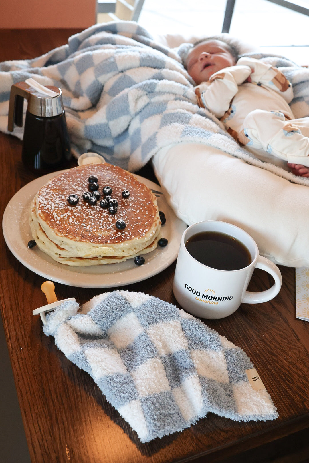 A cozy breakfast scene features a stack of pancakes with blueberries, coffee, syrup, and a soft Morning Check blanket. A sleeping baby cuddles the Lovey | Morning Check by forever french baby at the table.