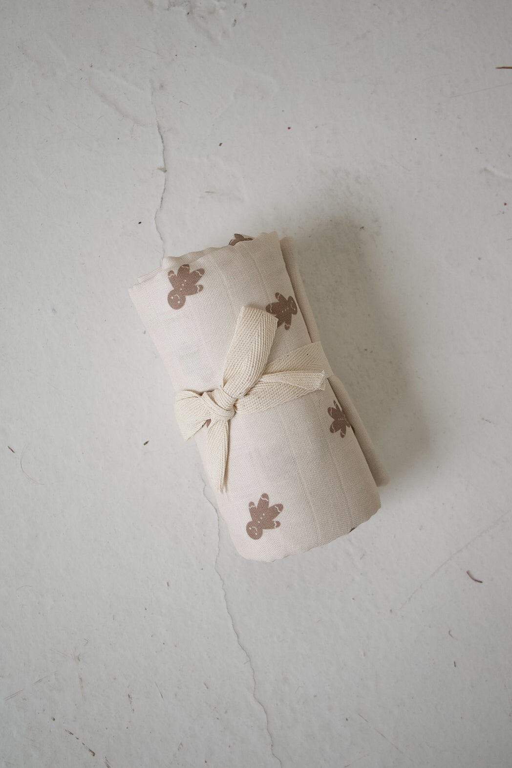 Forever French Baby Gingersnap muslin swaddle in cream cotton, featuring a brown gingerbread man print and tied with a matching fabric ribbon.