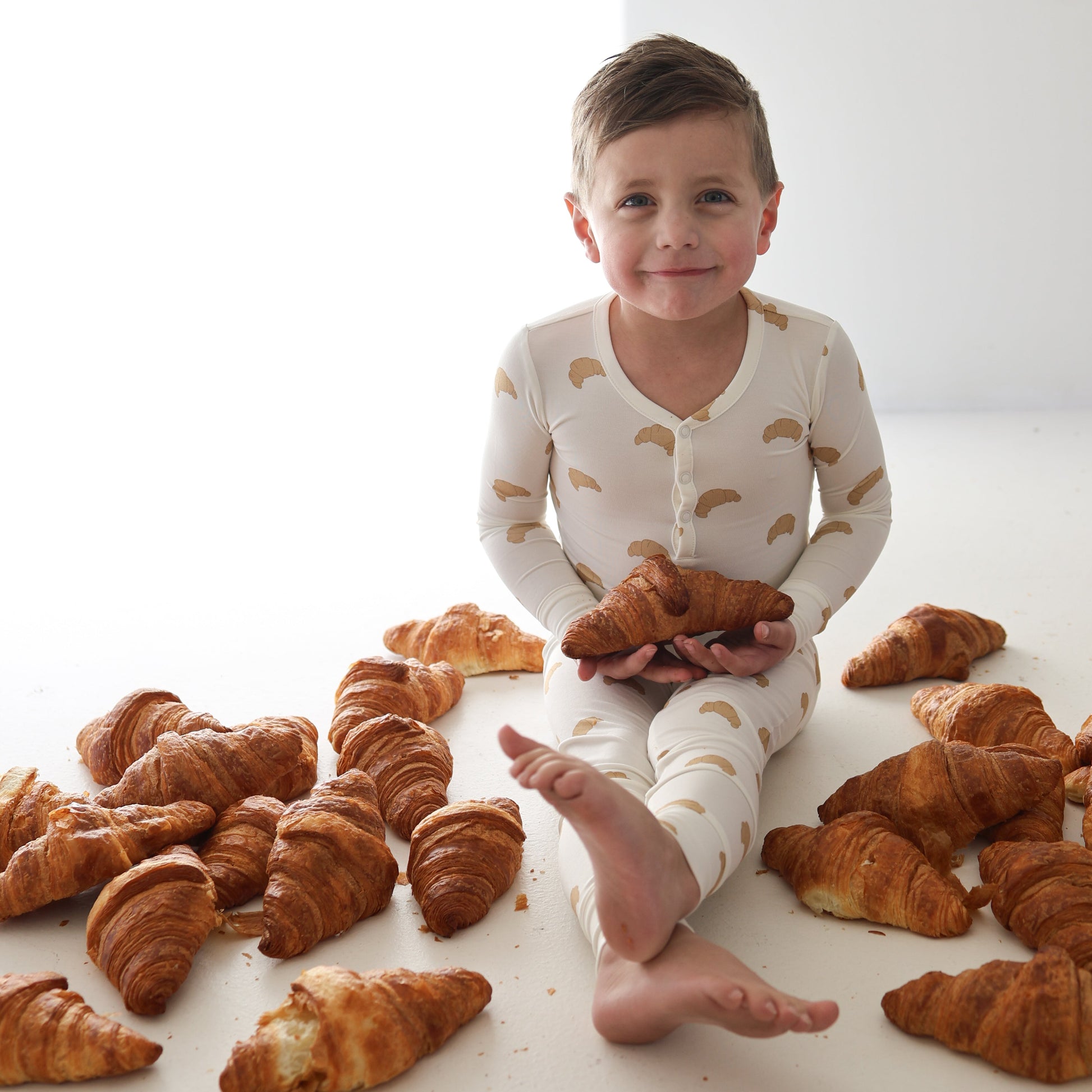 A young child wearing forever french baby's Bamboo Two Piece Pajamas in Croissants print sits on the floor, smiling and holding a croissant, with more croissants scattered around them against a white background.