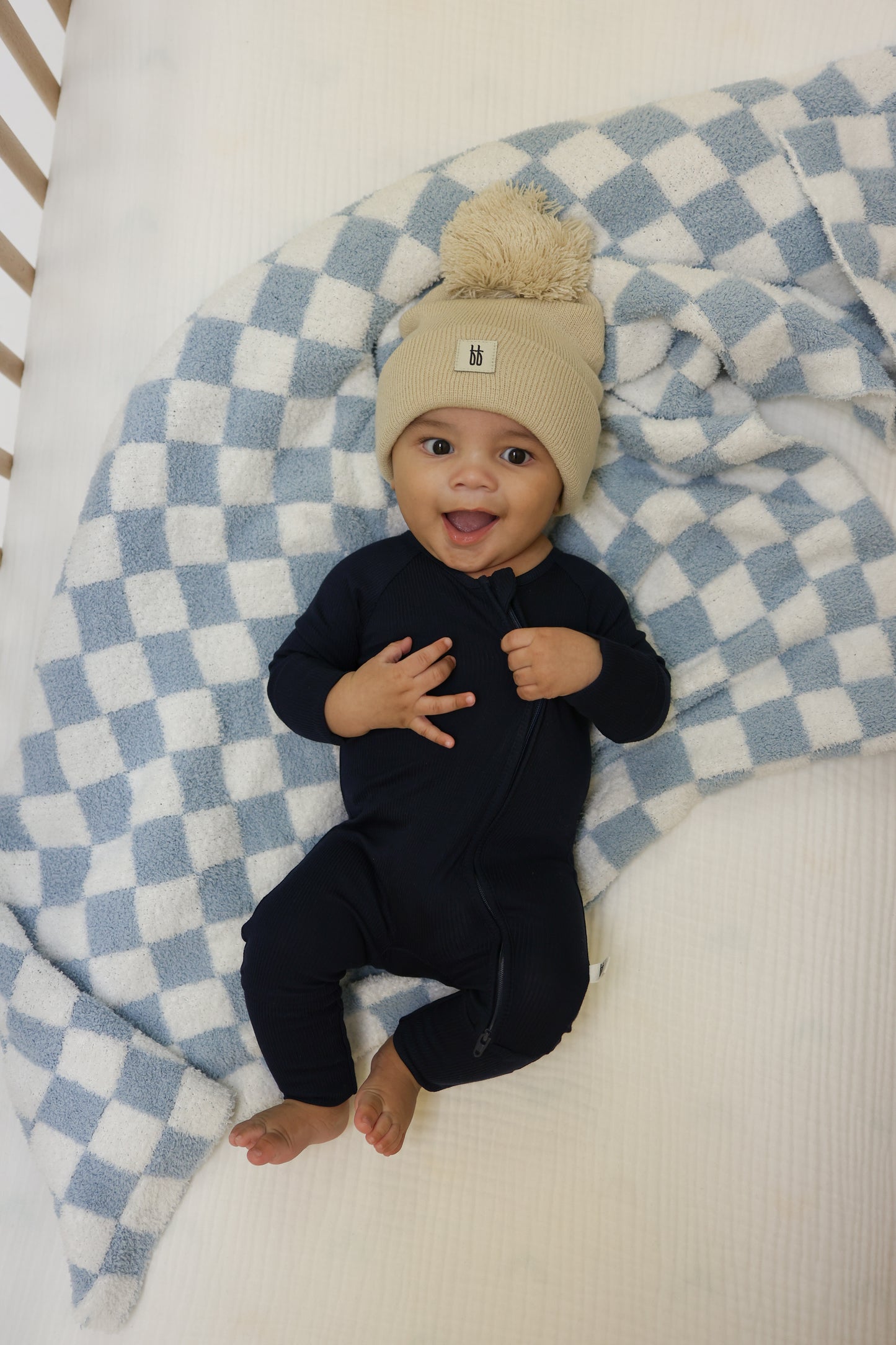 A smiling baby in forever french baby Bamboo Zip Pajamas | Blueberry Ribbed and a beige knit pom-pom hat lies on a light blue and white checkered blanket in a crib.