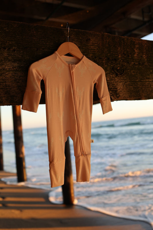 The forever french baby Bamboo Zip Pajamas | Lobster, a hypoallergenic long-sleeve and long-leg onesie in light pink, hangs on a wooden hanger beneath a pier with sunset waves and sandy beach in the background.