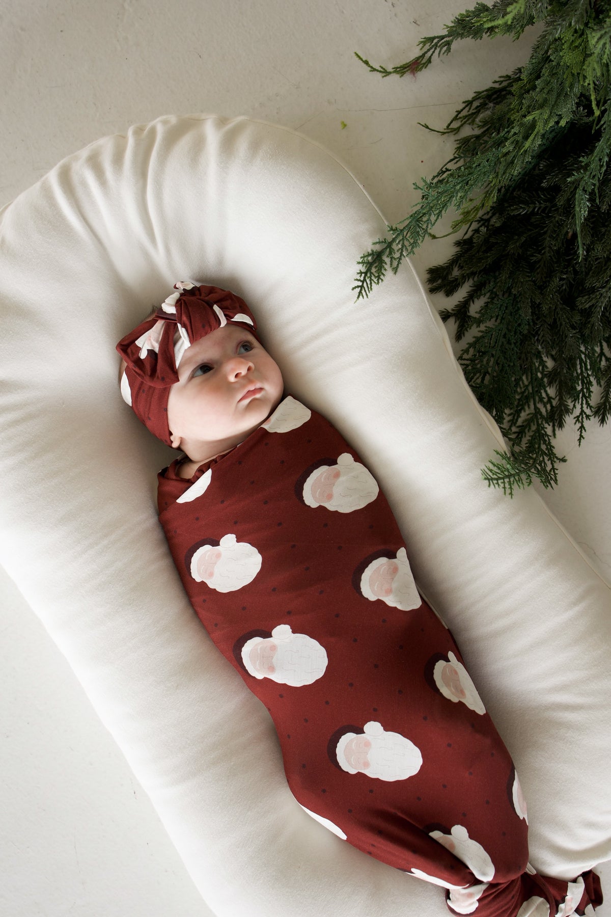 A baby in a Bamboo Swaddle | Vintage Santa and matching knotted headband, resting in a cream-colored baby lounger.