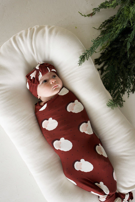 A baby in a Bamboo Swaddle | Vintage Santa and matching knotted headband, resting in a cream-colored baby lounger.