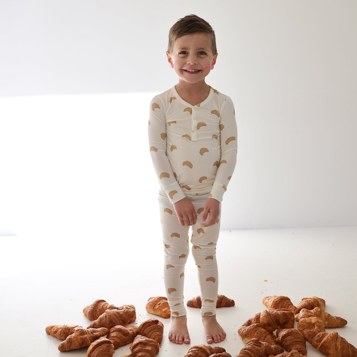 A smiling child stands barefoot on a white floor with croissants, wearing forever french baby's Bamboo Two Piece Pajamas | Croissants—hypo-allergenic, cream-colored sleepwear that's perfect for comfort in any well-lit, plain room.