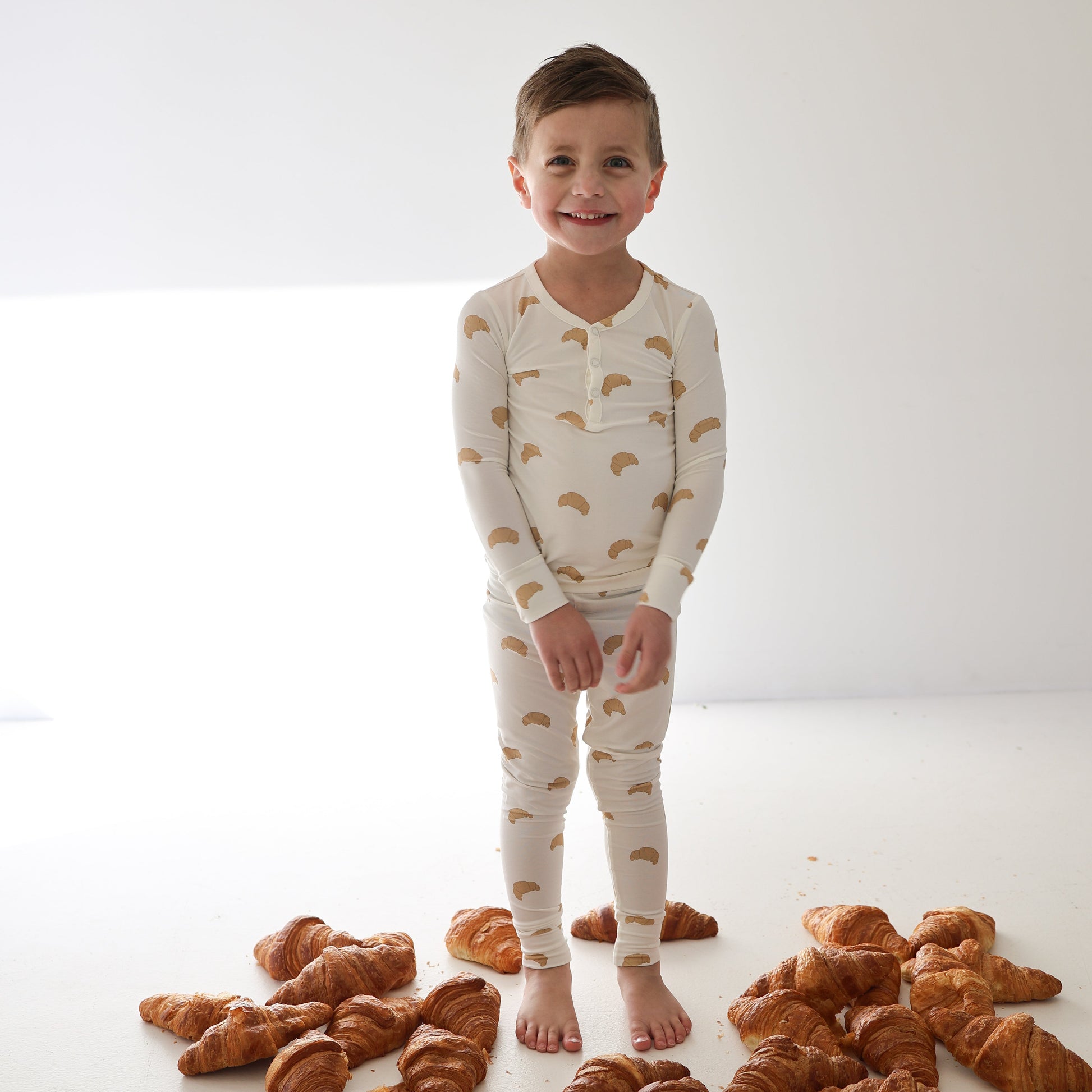 A smiling child stands barefoot on a white floor with croissants, wearing forever french baby's Bamboo Two Piece Pajamas | Croissants—hypo-allergenic, cream-colored sleepwear that's perfect for comfort in any well-lit, plain room.