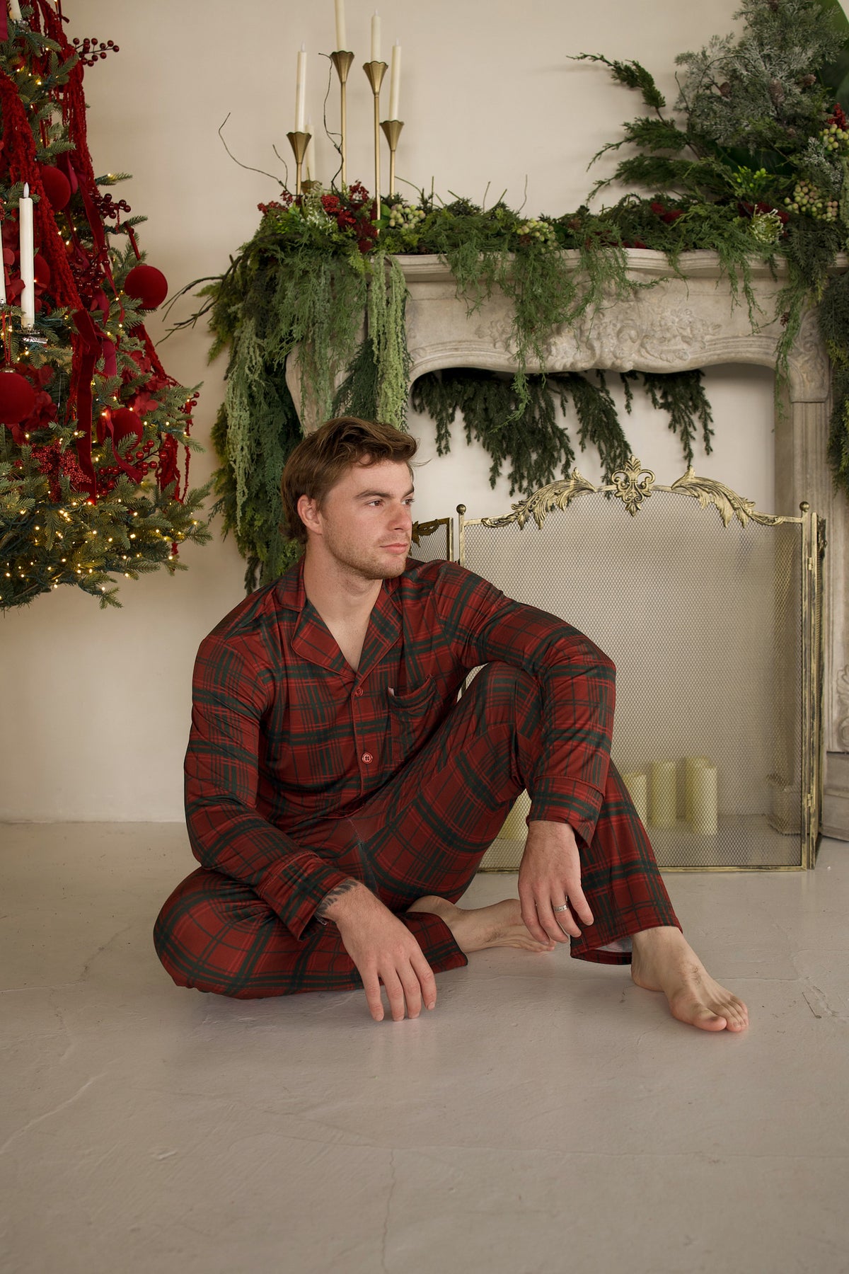 A man sits on the floor wearing a forever french baby Men’s Bamboo Button Up Lounge Set in Christmas Plaid in front of a fireplace and Christmas tree.