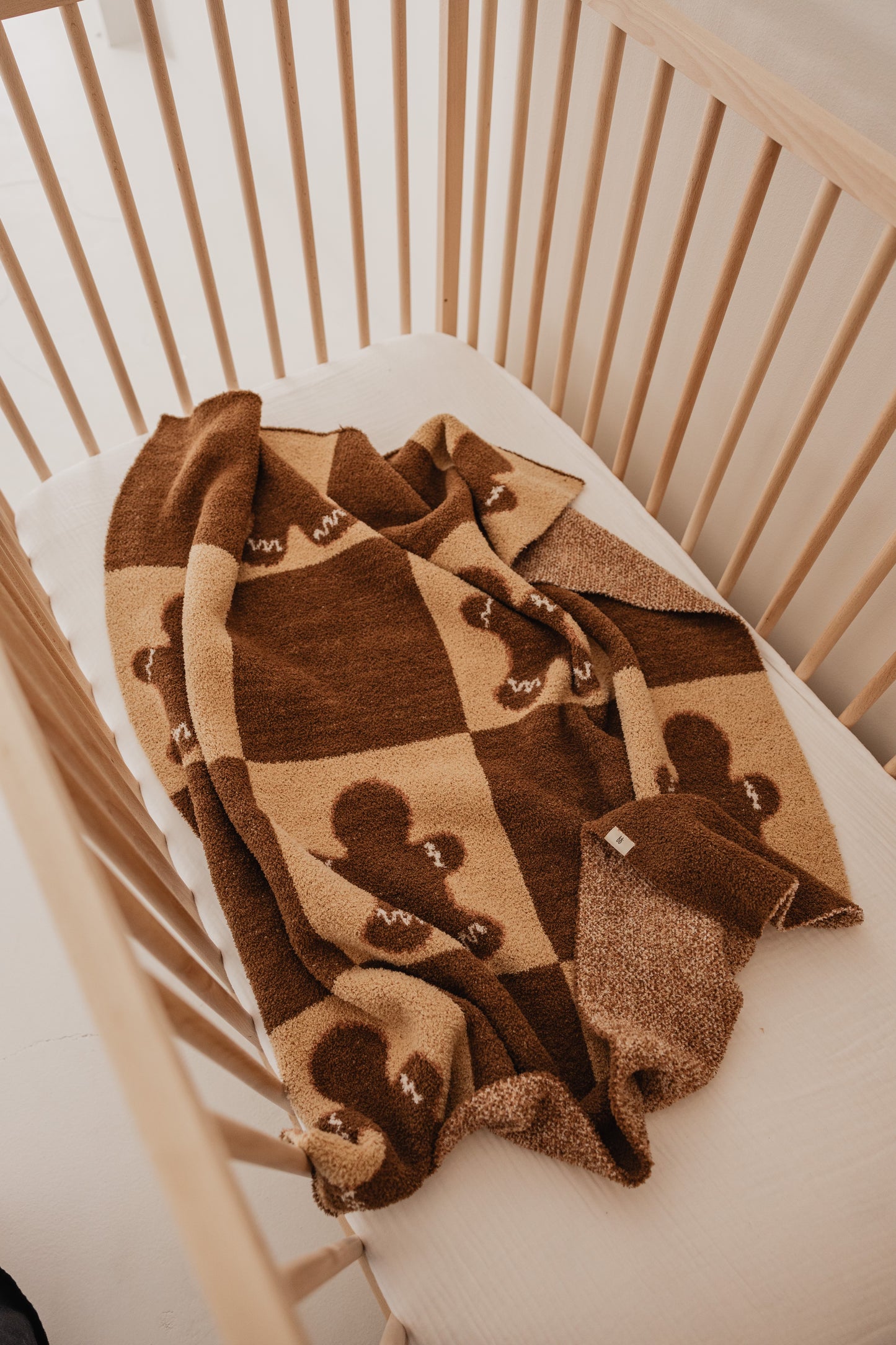 A wooden crib with a beige mattress is complemented by the forever french baby Gingerbread Checkerboard Plush Blanket. This cozy, minimalist setting highlights the blanket's soft brown and tan microfiber feather yarn, adorned with teddy bear and letter "M" motifs, all crafted to OEKO Standard 100.