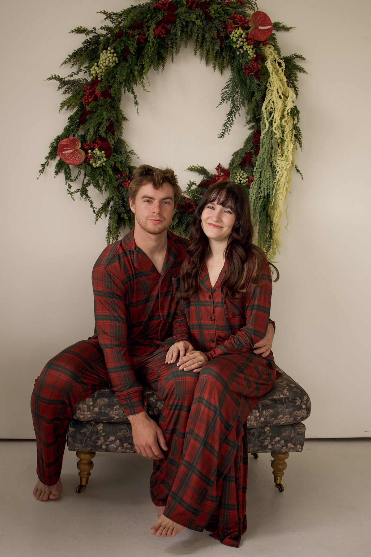 A man and a woman in matching Christmas Plaid Men’s Bamboo Button Up Lounge Set pajamas sit on an upholstered bench in front of a large holiday wreath.