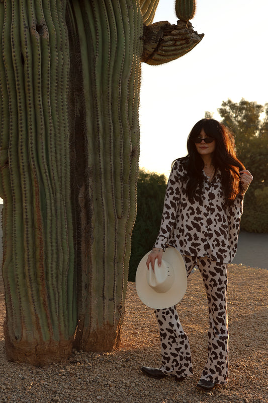 A woman in the forever french baby Women's Bamboo Button Up Lounge Set | Moo! and holding a white hat stands next to a cactus in the desert at sunset.