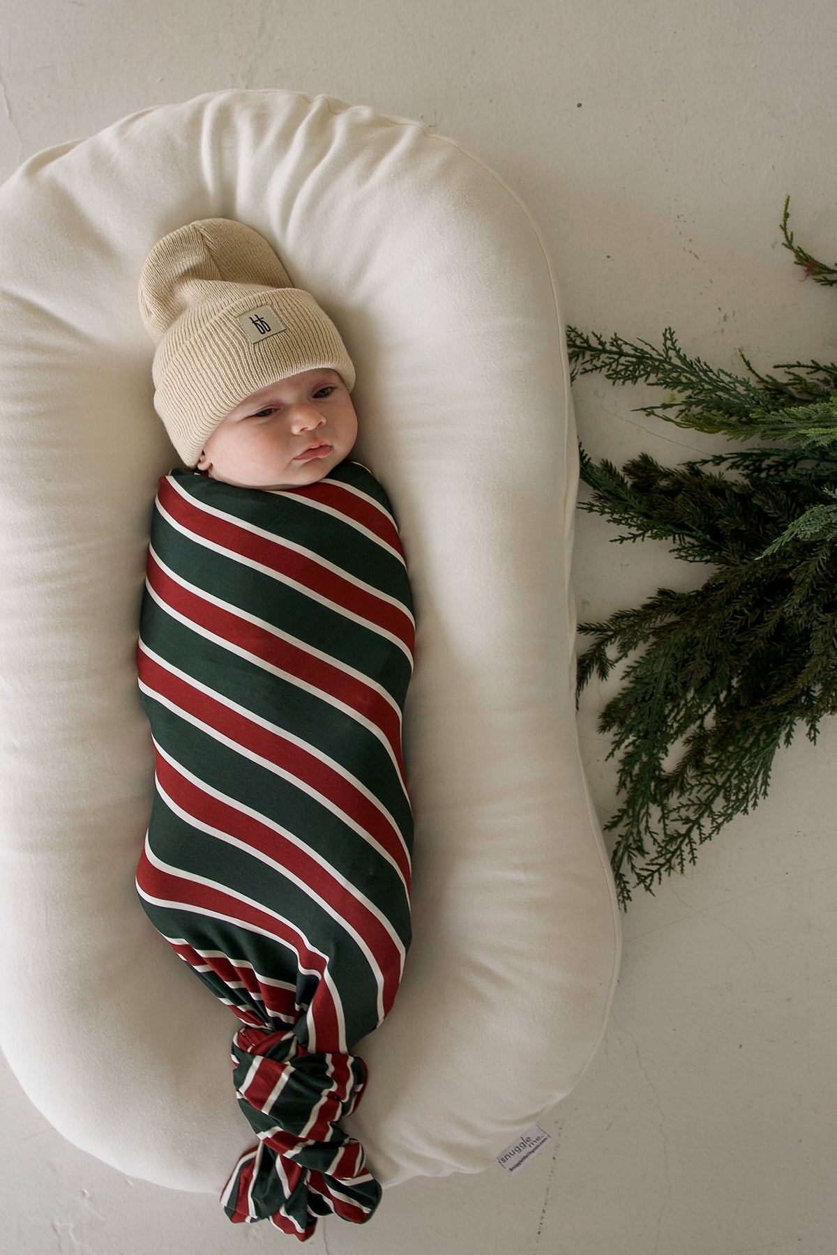 A baby wearing a tan beanie lies in a crescent-shaped lounger, wrapped in a Bamboo Swaddle | Holiday Stripe.