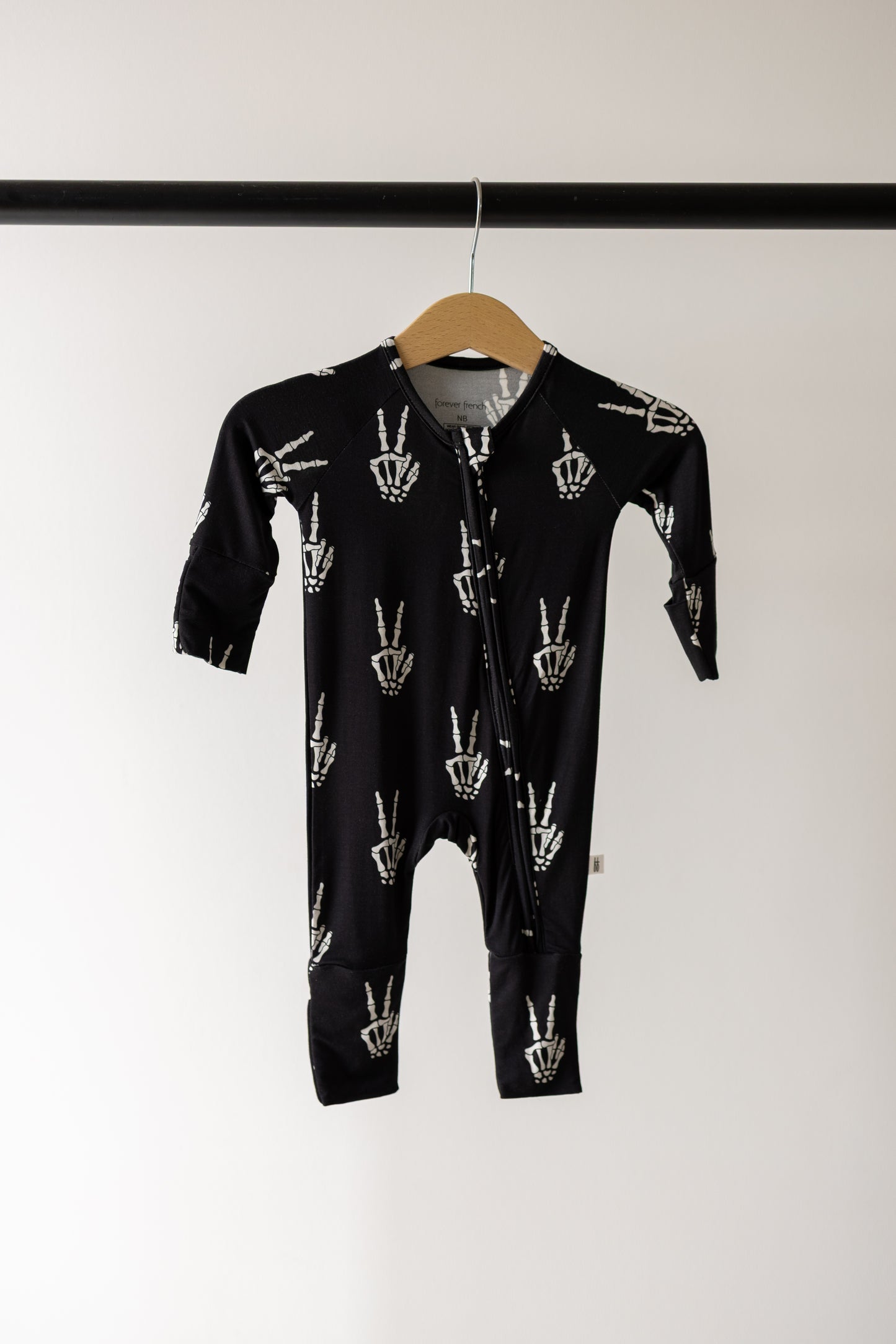 The Bamboo Zip Pajamas | Midnight Skelly by forever french baby feature a black design with white hand skeletons showing peace signs, crafted from ultra-soft bamboo viscose for exceptional breathability and comfort. Hanging on a wooden hanger against a plain background, these hypo-allergenic pajamas are an ideal choice for babies with sensitive skin.