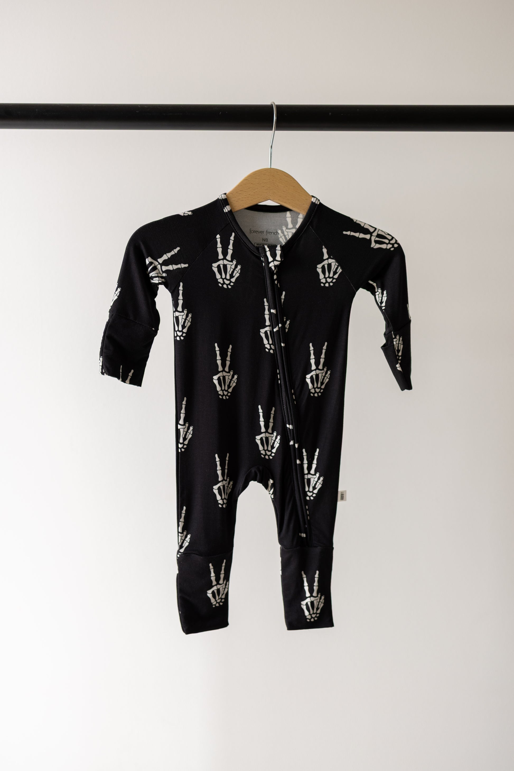 The Bamboo Zip Pajamas | Midnight Skelly by forever french baby feature a black design with white hand skeletons showing peace signs, crafted from ultra-soft bamboo viscose for exceptional breathability and comfort. Hanging on a wooden hanger against a plain background, these hypo-allergenic pajamas are an ideal choice for babies with sensitive skin.