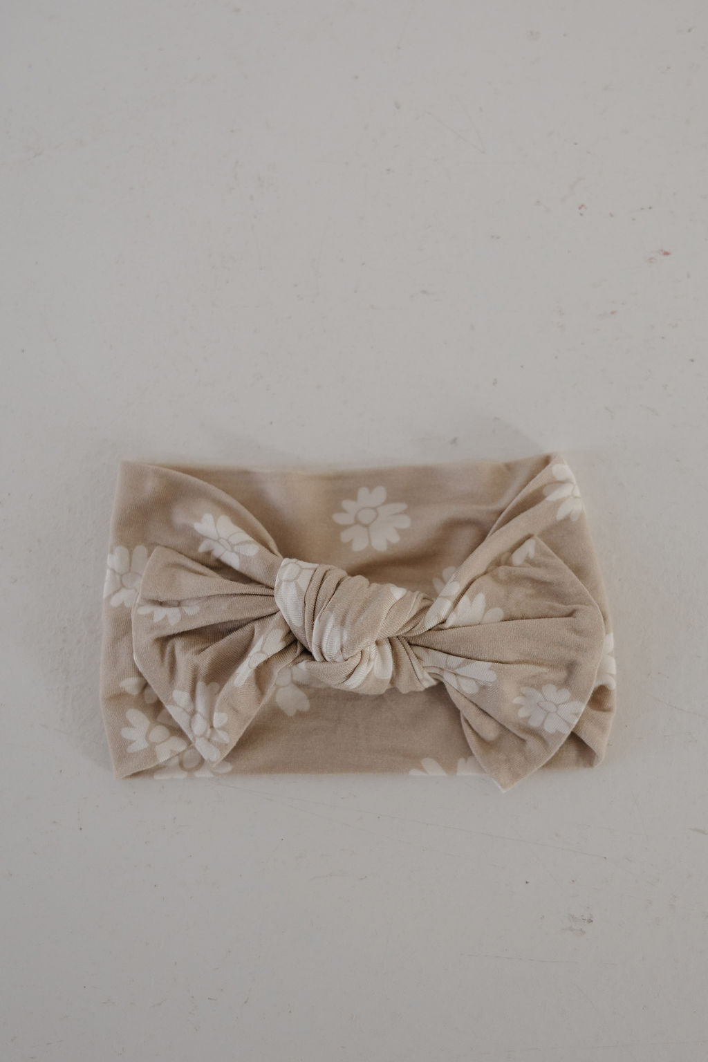 The Bamboo Head Wrap | Lazy Daisy by forever french baby, featuring white floral patterns and a large central knot in beige, is displayed on a light gray surface—an adorable accessory for any outfit.