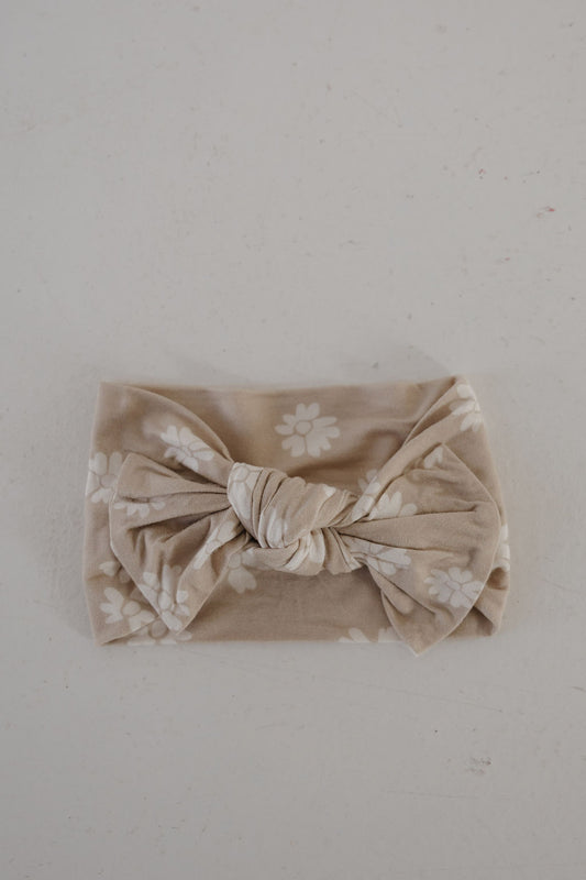 The Bamboo Head Wrap | Lazy Daisy by forever french baby, featuring white floral patterns and a large central knot in beige, is displayed on a light gray surface—an adorable accessory for any outfit.