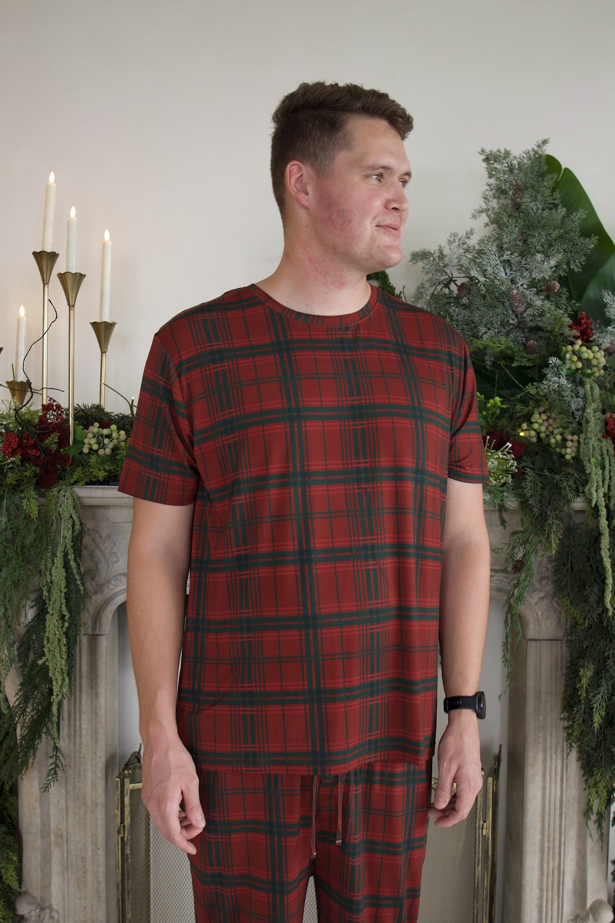 A man wears the Men’s Short Sleeve Pajamas | Christmas Plaid, featuring a short-sleeve crewneck shirt and matching pants in a red and green plaid pattern.