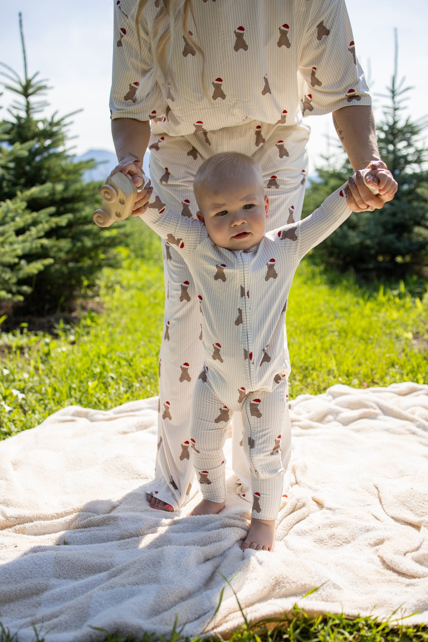 Clad in forever french baby’s Bamboo Zip Pajamas | Santa Teddy, a baby with matching festive prints is held up by an adult, standing on a blanket outdoors among pine trees on a sunny day.