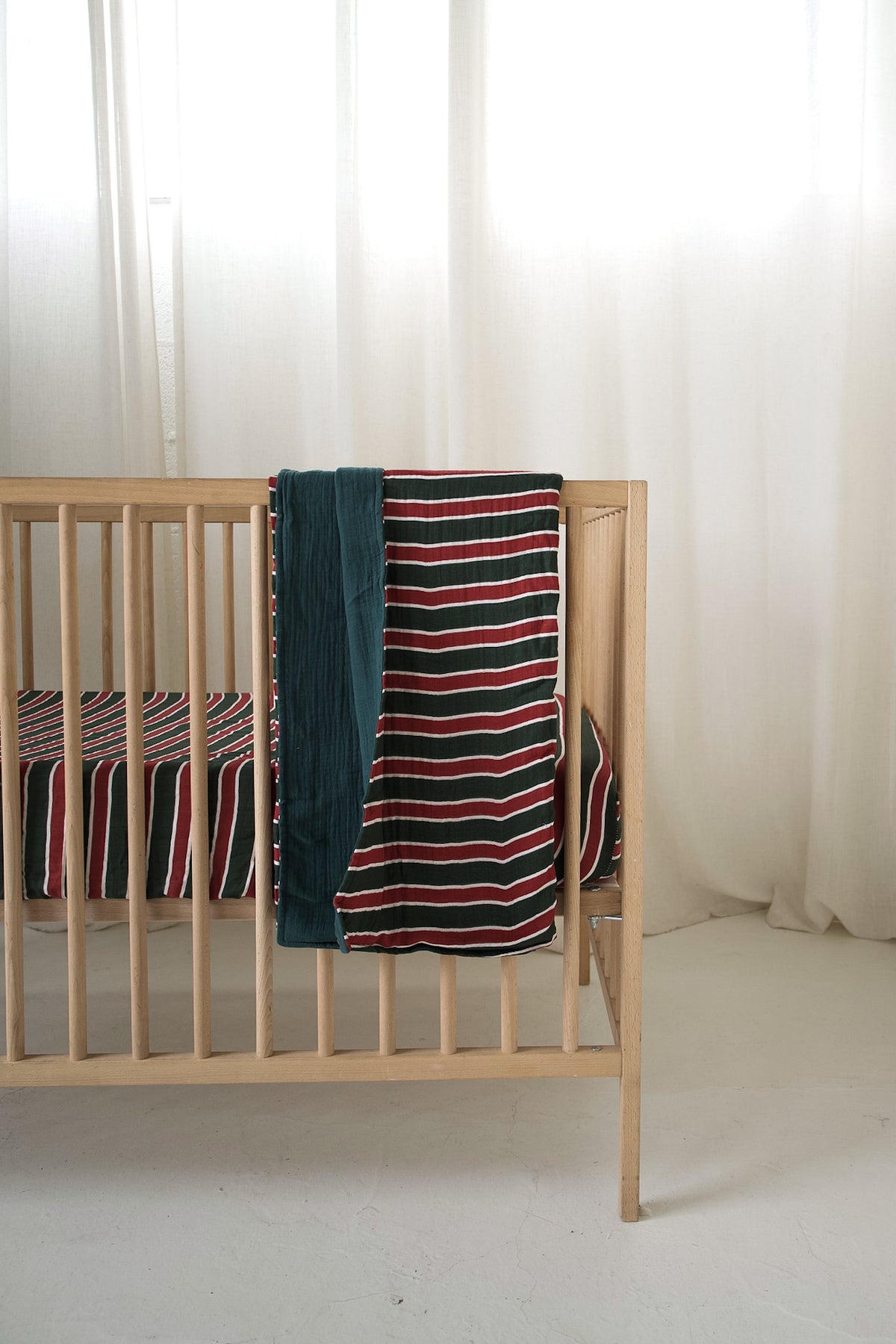A wooden crib draped with a forever french baby Holiday Stripe muslin quilt in red, white, and green.