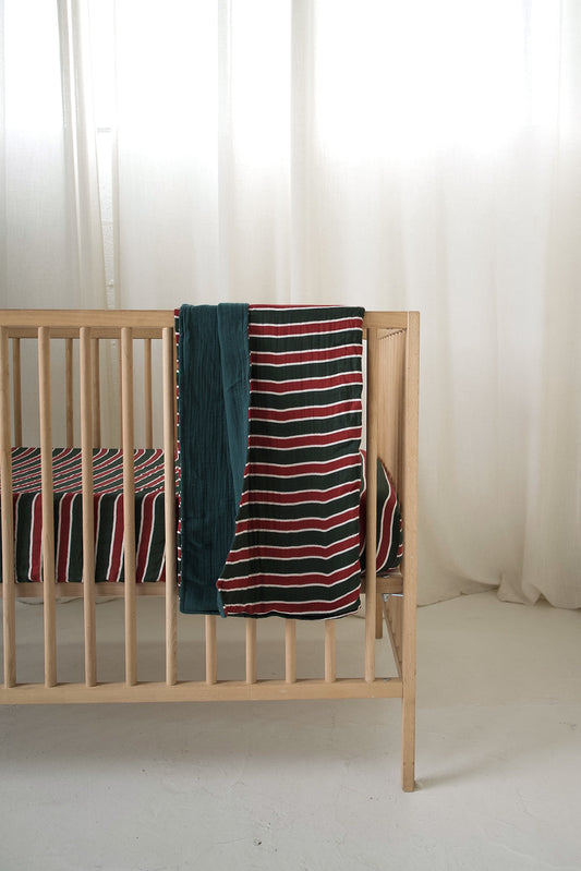 A wooden crib draped with a forever french baby Holiday Stripe muslin quilt in red, white, and green.