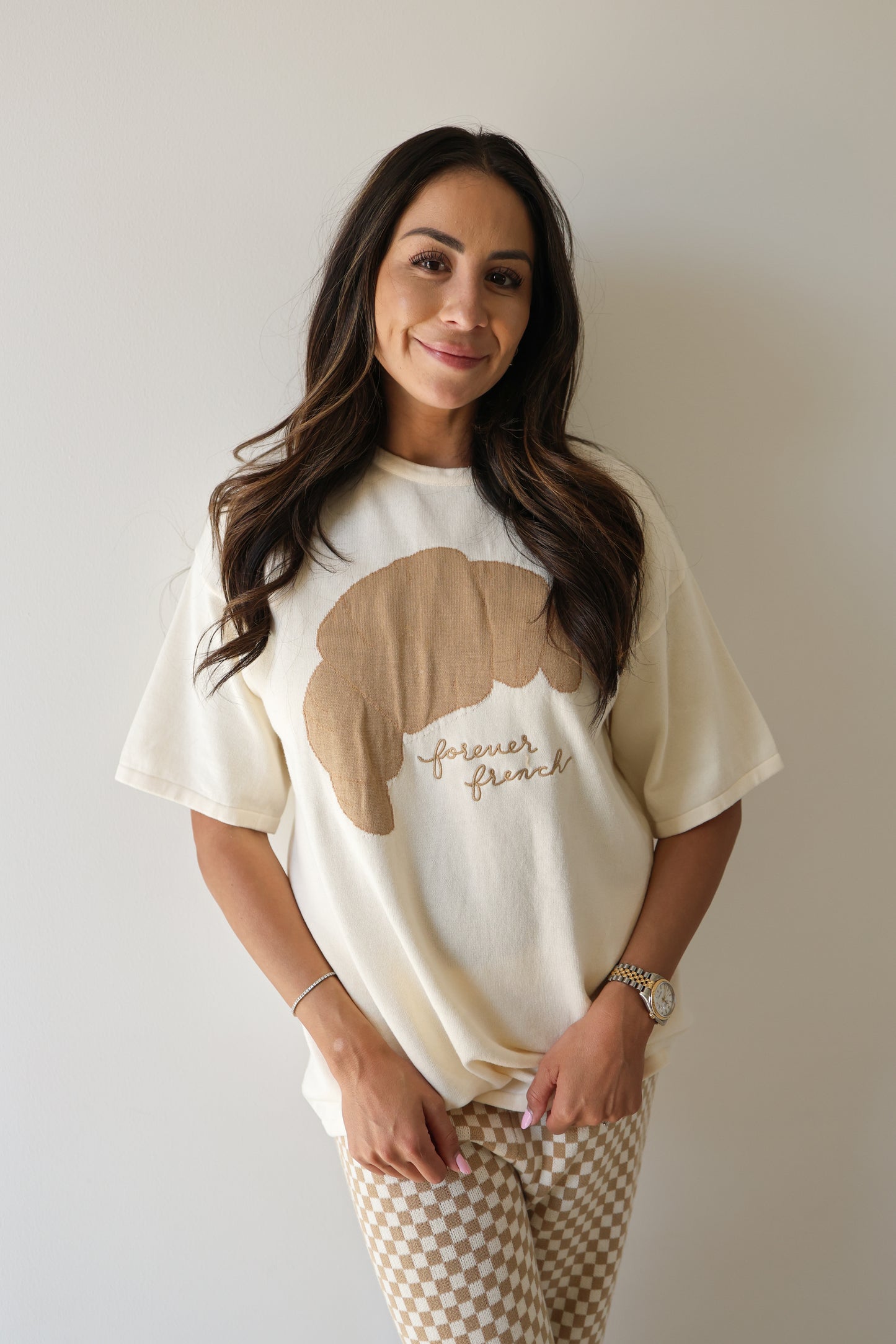 A woman with long dark hair stands against a light wall, smiling. She wears the Women's Knit Pant Set | Croissants by forever french baby, featuring a croissant graphic and checkered beige and white pants.
