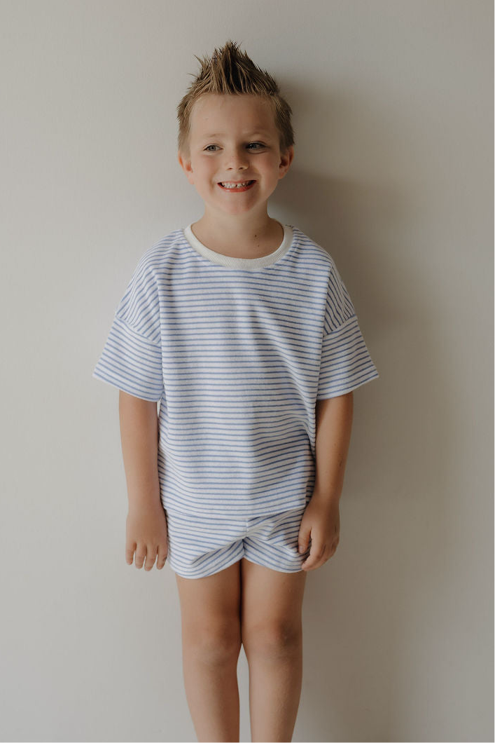 A young boy with short, spiked hair smiles at the camera against a light wall, wearing the forever french baby Child Terry Short Set in Ocean Stripe—perfect for capturing sweet family moments.