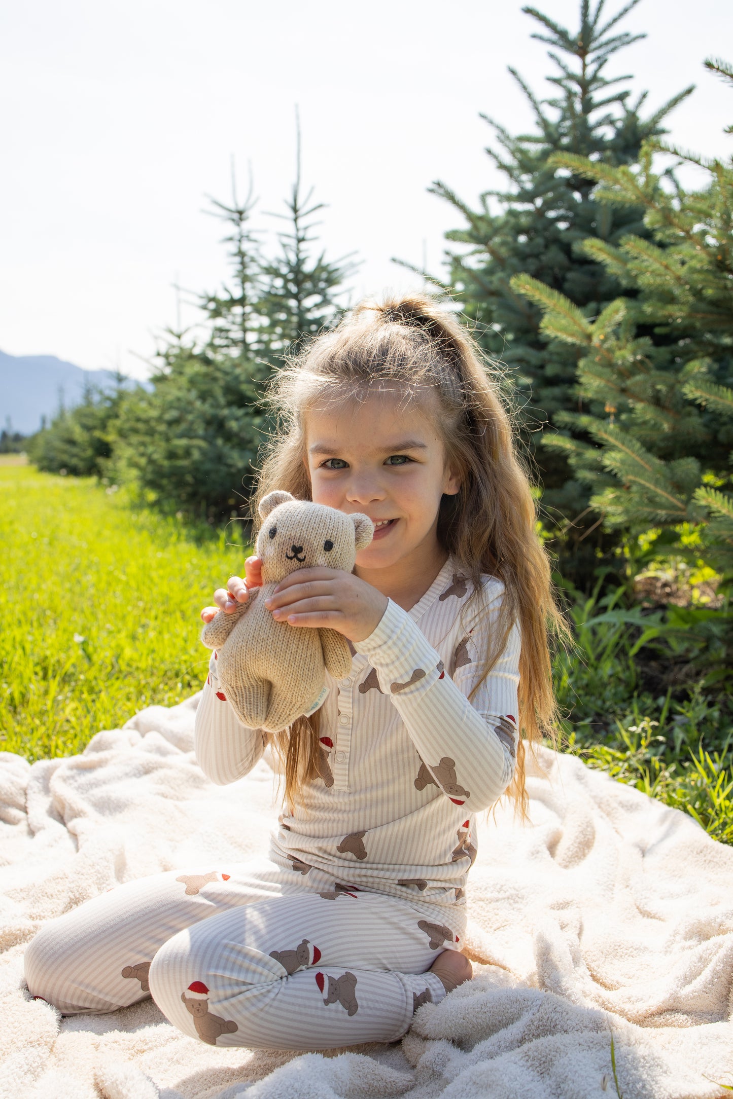 A young girl, smiling in her forever french baby Bamboo Two Piece Pajamas | Santa Teddy, sits on a blanket outdoors surrounded by green pine trees on a sunny day.