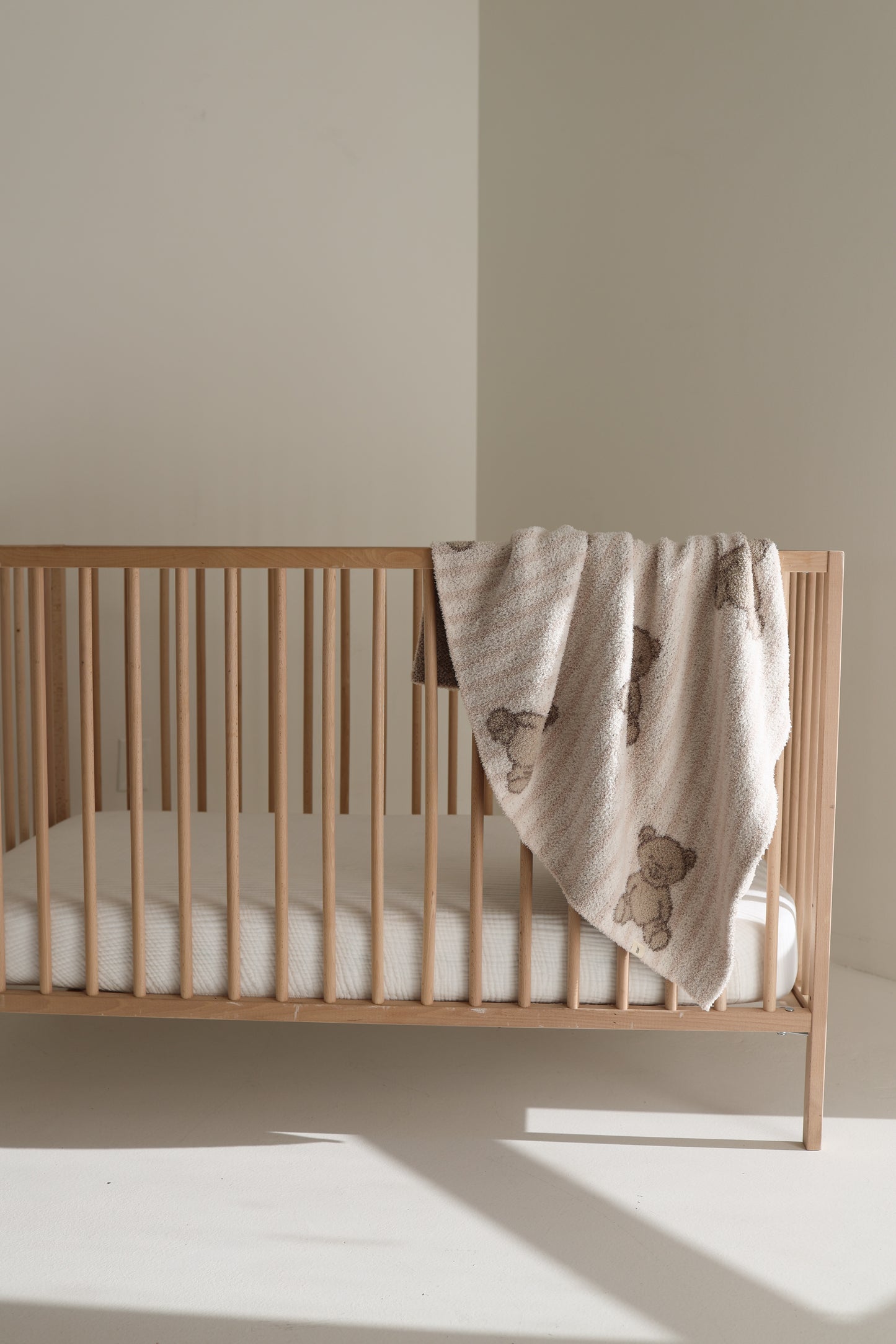 A wooden baby crib with a white mattress stands in a bright, minimal room. A forever french baby Plush Blanket | Teddy—Oeko-Tex certified and adorned with teddy bear patterns—is draped over one side of the crib.