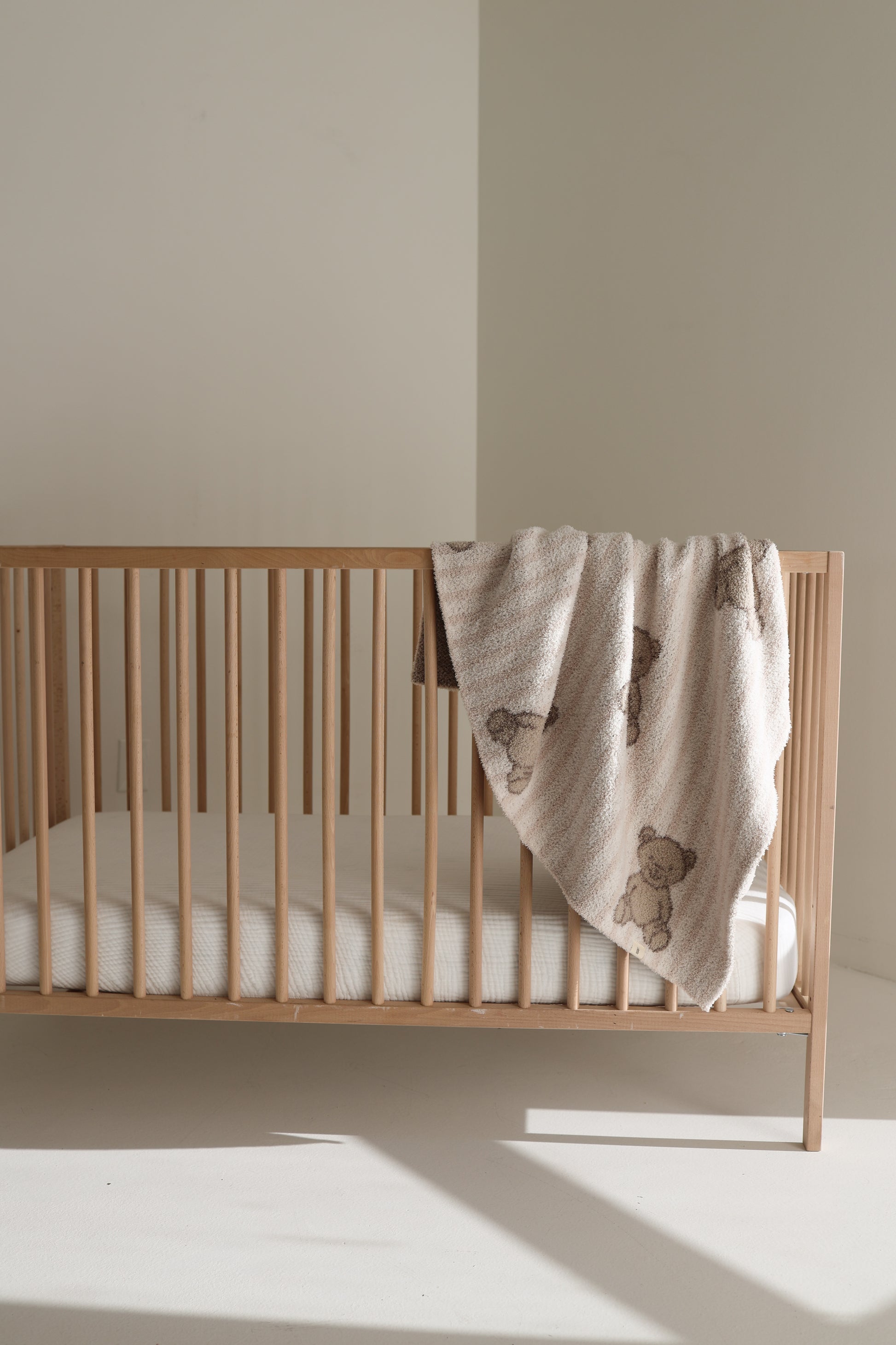A wooden baby crib with a white mattress stands in a bright, minimal room. A forever french baby Plush Blanket | Teddy—Oeko-Tex certified and adorned with teddy bear patterns—is draped over one side of the crib.