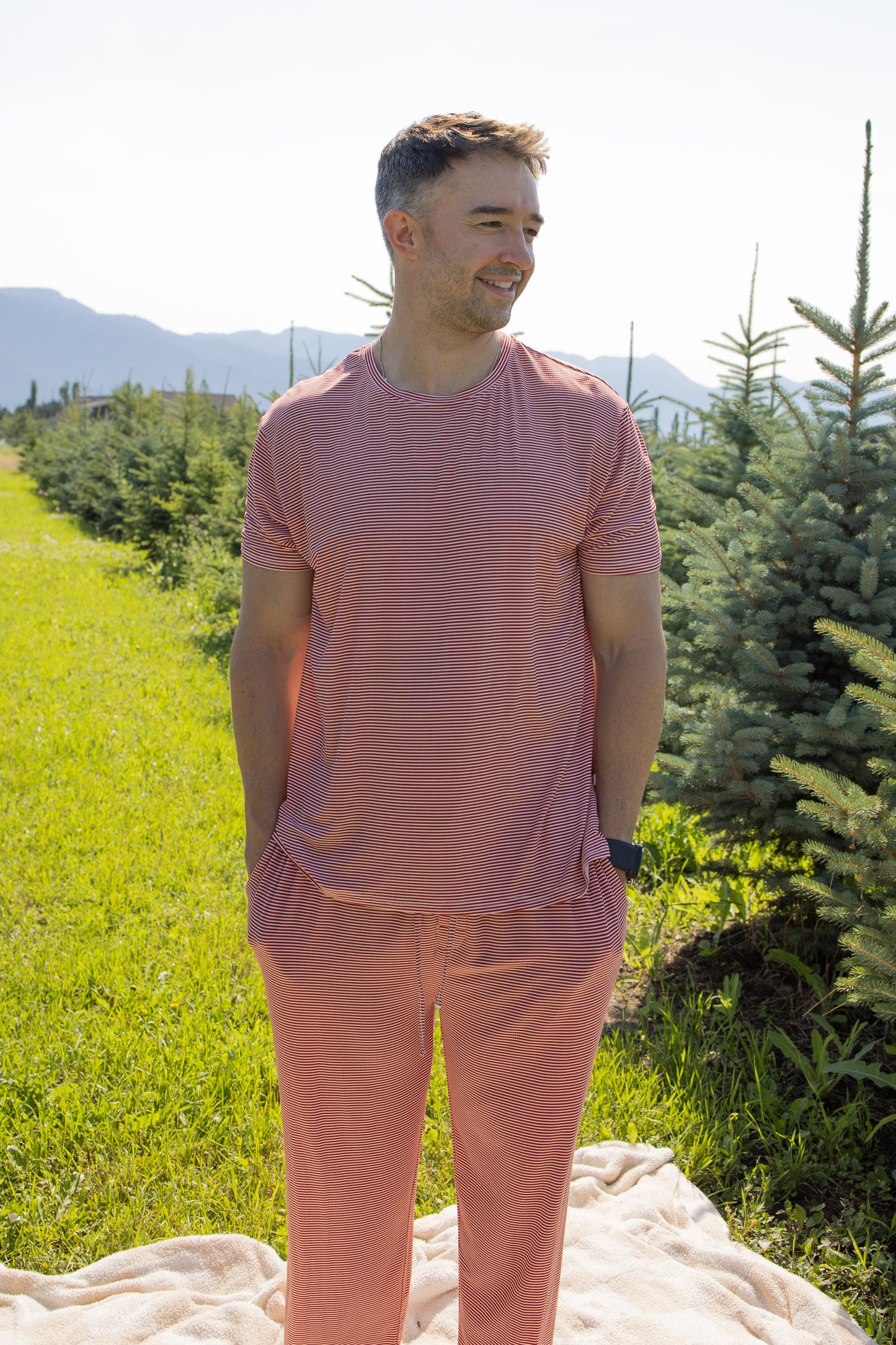 A man wearing the forever french baby Men's Short Sleeve Pajamas in Peppermint Stripe Ribbed stands on a blanket in a green field between rows of small pine trees, with mountains and a clear sky in the background.