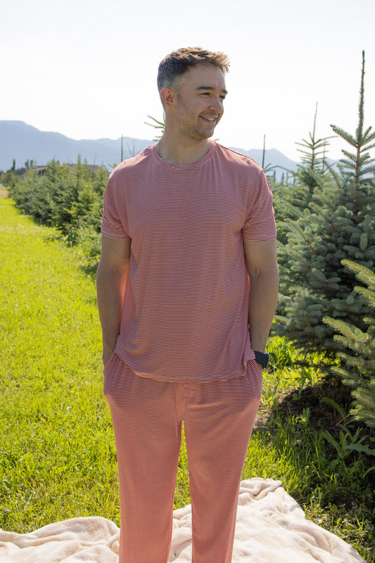 A man wearing the forever french baby Men's Short Sleeve Pajamas in Peppermint Stripe Ribbed stands on a blanket in a green field between rows of small pine trees, with mountains and a clear sky in the background.