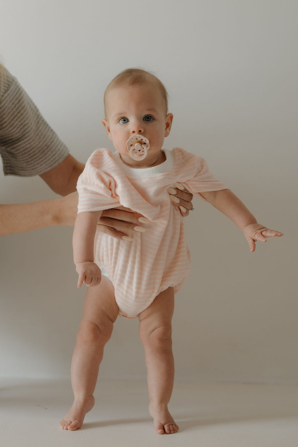 A baby wearing the forever french baby Terry T-Shirt Romper in Cotton Candy stands with support from an adult, pacifier in hand, capturing precious family moments against a plain light background.