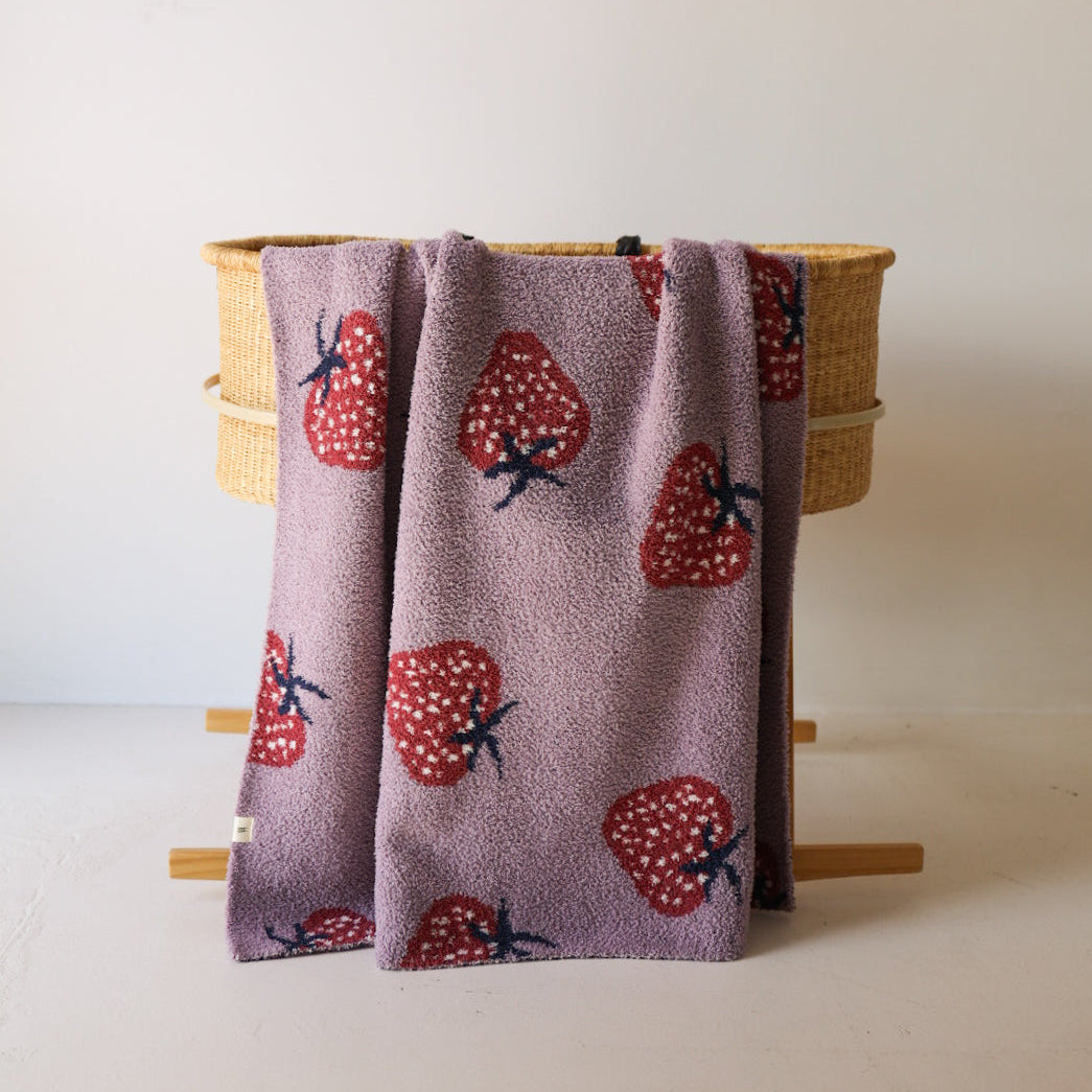 The forever french baby Plush Blanket | Matilda, in lavender velvet with red and white strawberry patterns, is draped over a woven basket with wooden legs against a plain white wall.