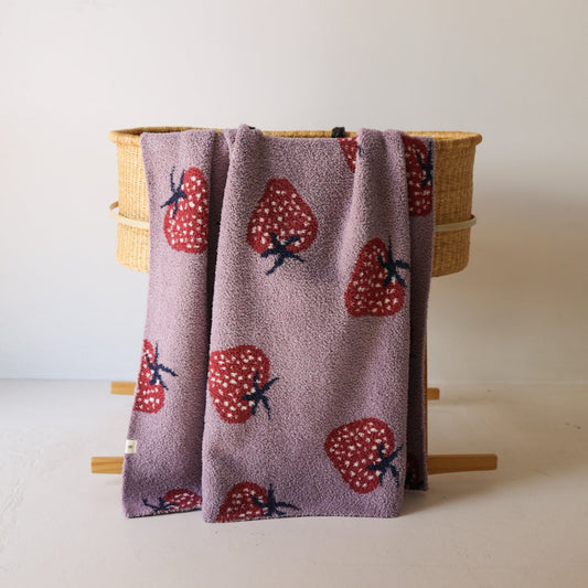The forever french baby Plush Blanket | Matilda, in lavender velvet with red and white strawberry patterns, is draped over a woven basket with wooden legs against a plain white wall.