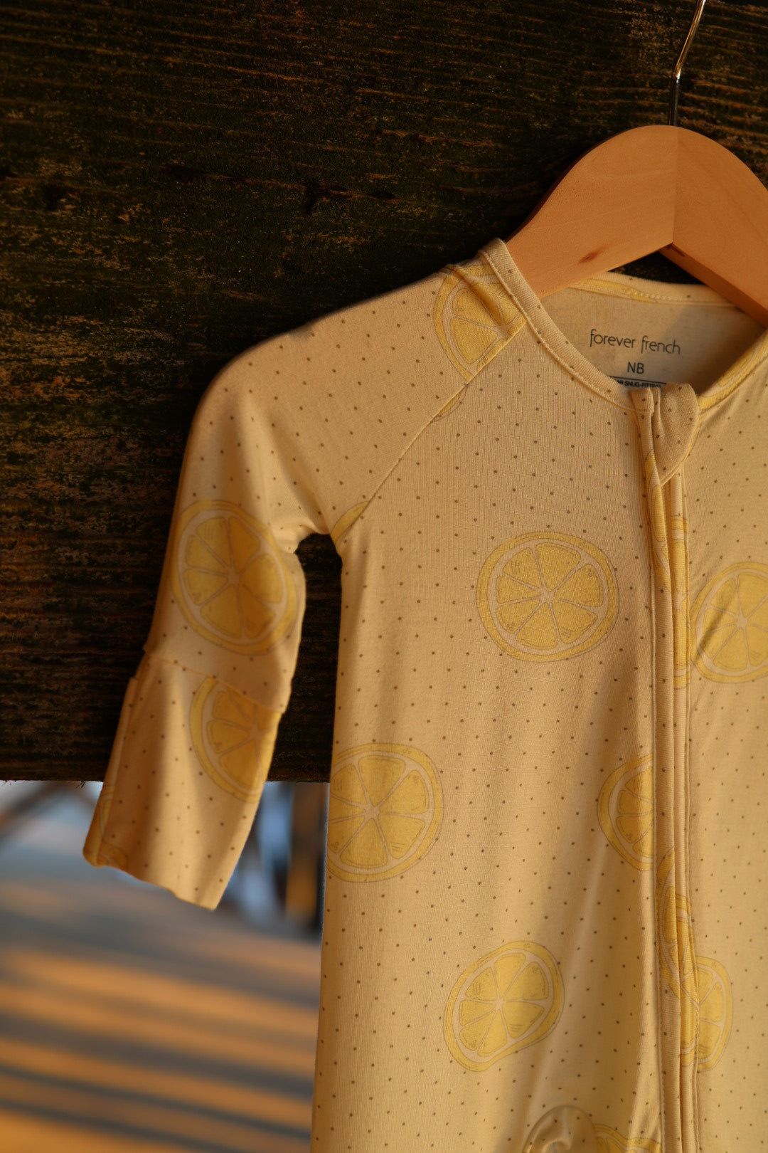 The "Bamboo Zip Pajamas | Lemon Drop" by forever french baby—a yellow, breathable baby onesie with a dotted pattern and lemon slice prints—hangs on a wooden hanger against a rustic background, bathed in warm sunlight.