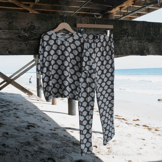 The forever french baby Child Boxy Tee Lounge Set in the Seashells print hangs on wooden hangers under a sunny beach pier, with ocean and sand in the background.