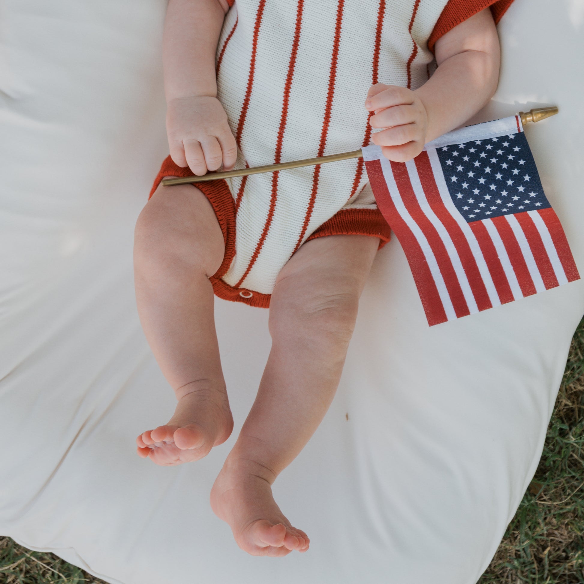 A baby wearing the forever french baby Knit Romper | Picnic Pinstripe sits outdoors on a white cushion, holding a small American flag. Only the baby's legs, arms, and flag-holding hand are visible.
