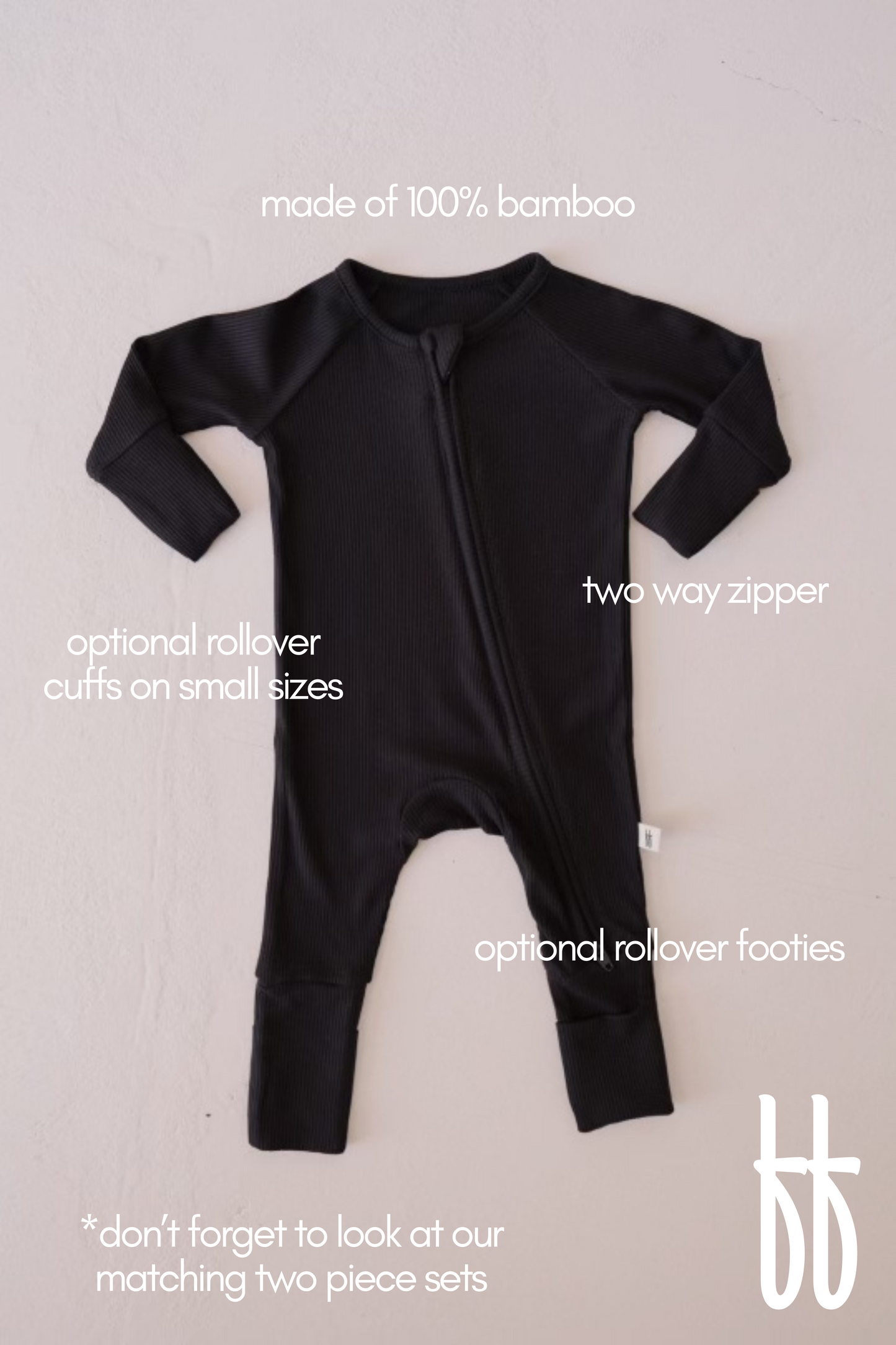 The Bamboo Zip Pajamas | Black Ribbed from forever french baby is displayed lying flat. Made from 100% bamboo, this hypo-allergenic baby onesie features a two-way zipper, optional rollover cuffs on smaller sizes, and optional rollover footies. Text on the image highlights these features, along with a note suggesting to check out matching two-piece sets.