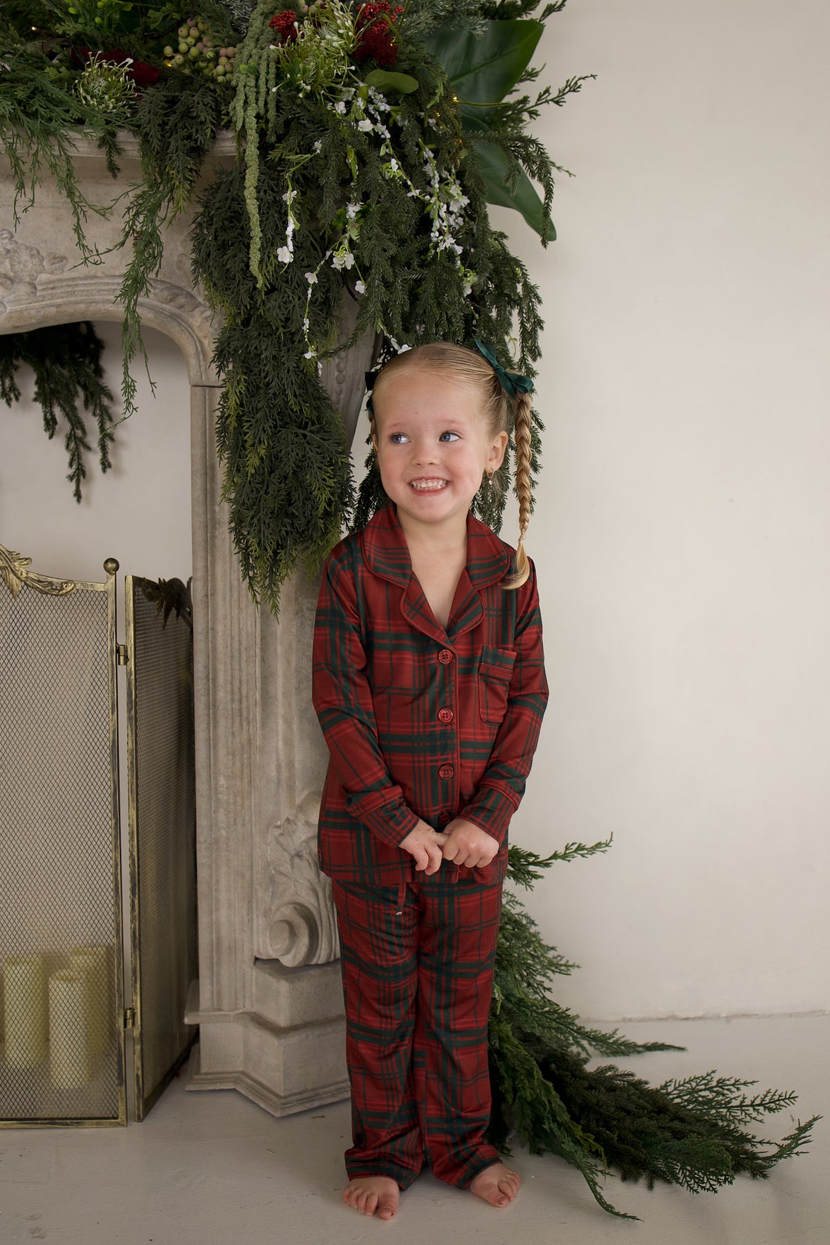 A smiling young girl stands by a fireplace wearing a Girl’s Bamboo Button Up Lounge Set in Christmas plaid, featuring a long-sleeved button-up top and matching pants.