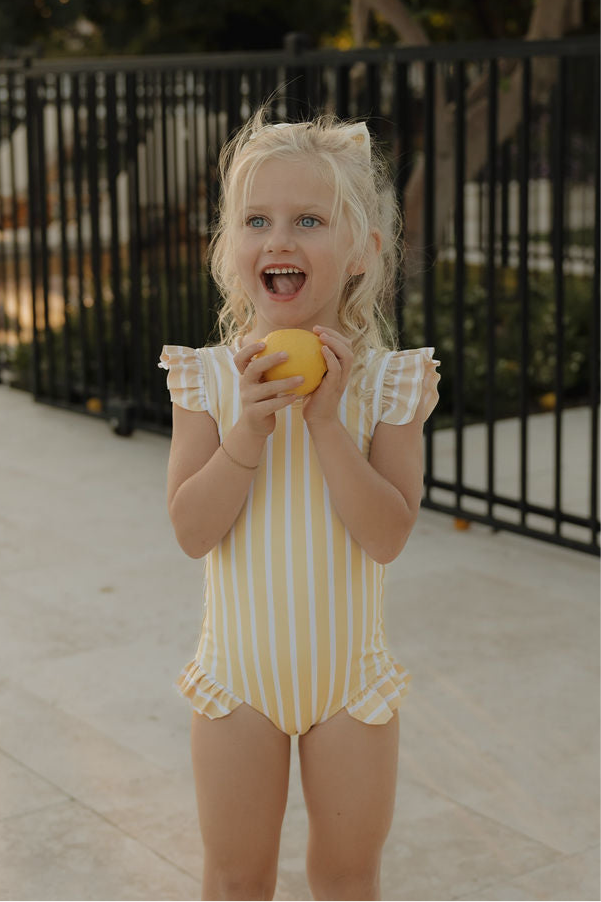 A young girl smiles outdoors in the lolo webb 2 Girl Sleeveless Swimsuit | Sun Rays, featuring yellow and white stripes with ruffle detail, holding a lemon by a black fence and greenery—capturing playful summer style.