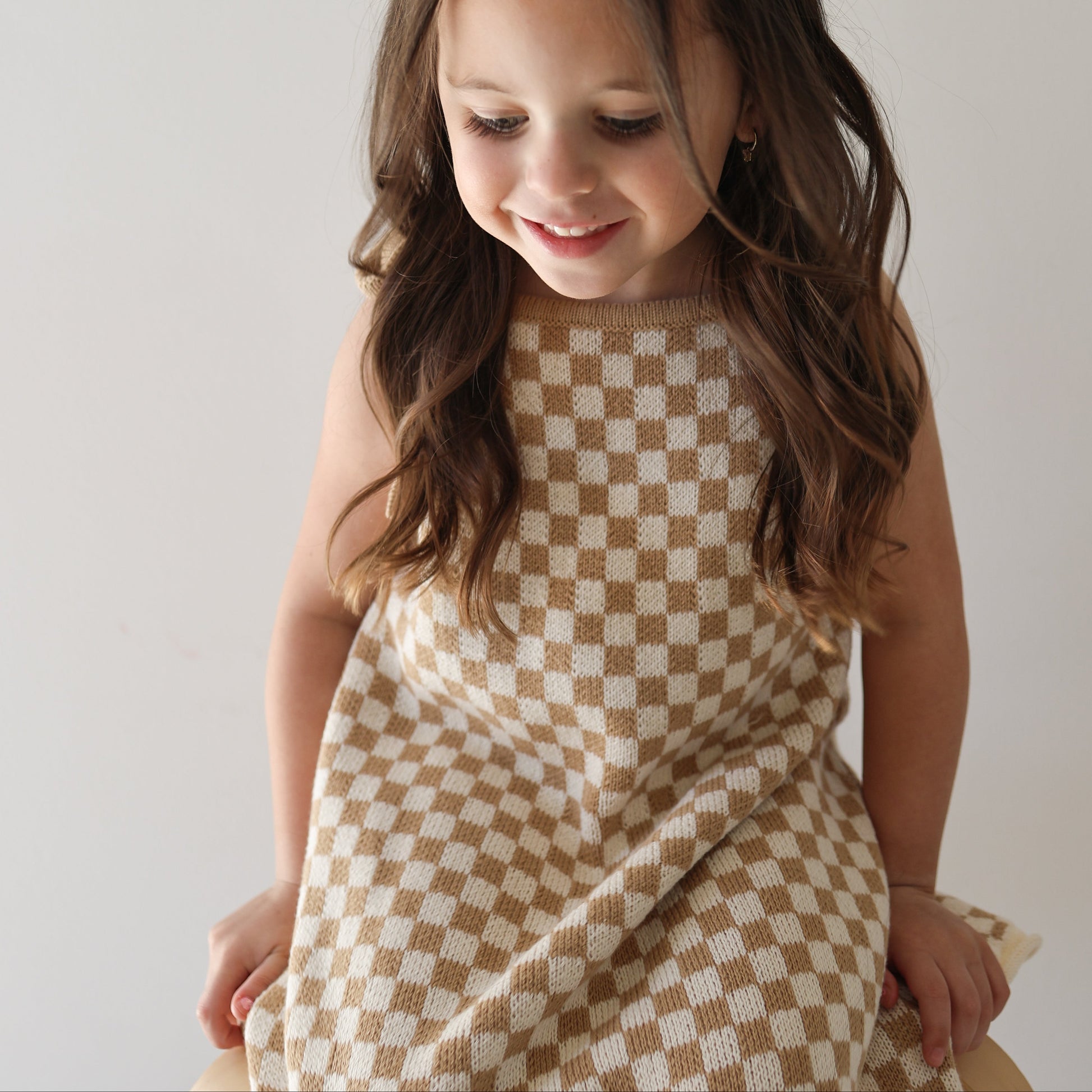 A young girl with long brown hair smiles while wearing the Knit Dress | Latte Check from forever french baby, seated against a plain light background.