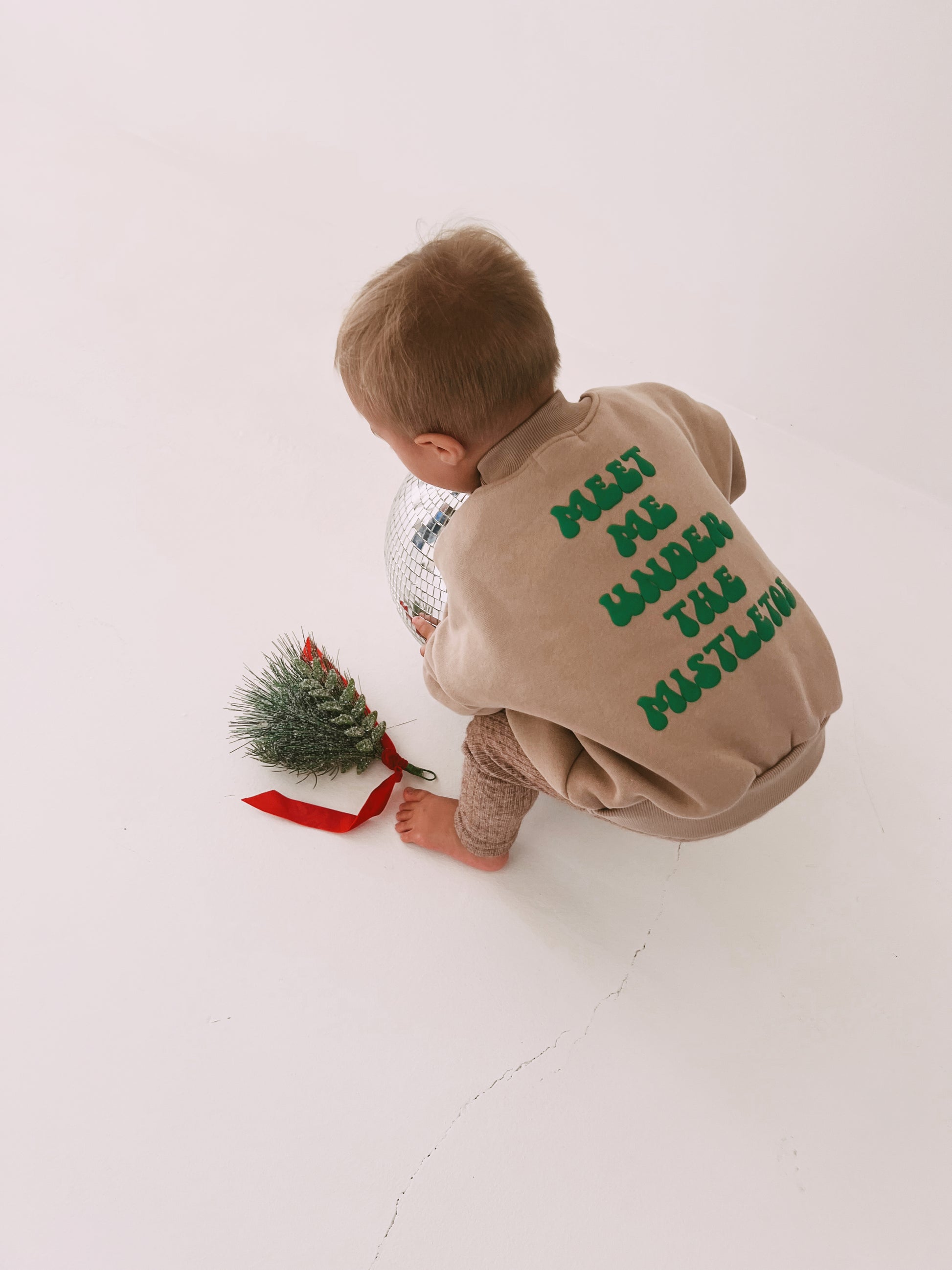 A child in a beige "Meet Me Under The Mistletoe" festive sweatshirt by forever french baby, featuring green text on the back, crouches on a white floor while holding a shiny silver ball. In front of the child lies a small artificial Christmas tree with a red base.