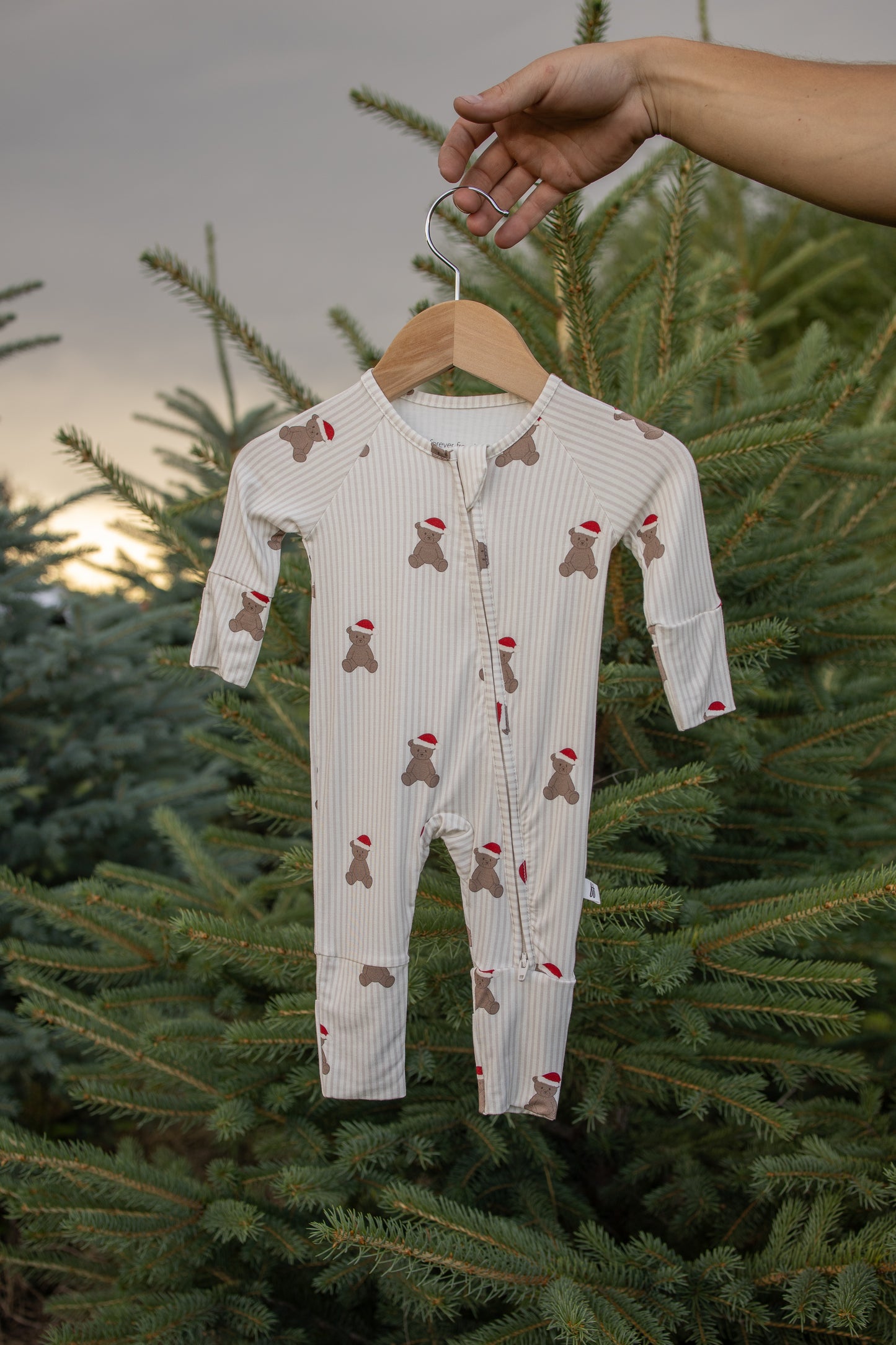 A hand holds a hanger with forever french baby's Bamboo Zip Pajamas | Santa Teddy, featuring gingerbread figures and tiny red hats, crafted from hypoallergenic materials, set against a backdrop of evergreen trees at sunset.