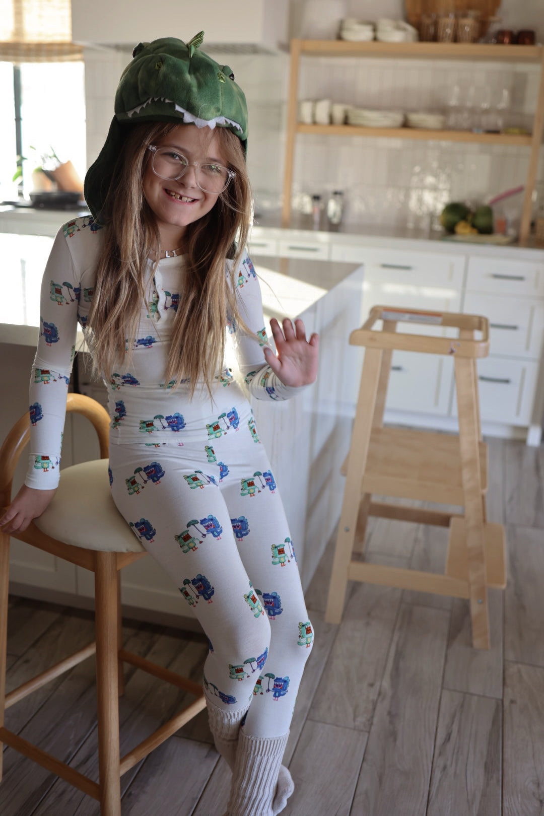 A young girl in glasses sits on a kitchen stool, smiling and waving at the camera while wearing forever french baby Bamboo Two Piece Pajamas | Charli's Monsters and a green dinosaur hat.