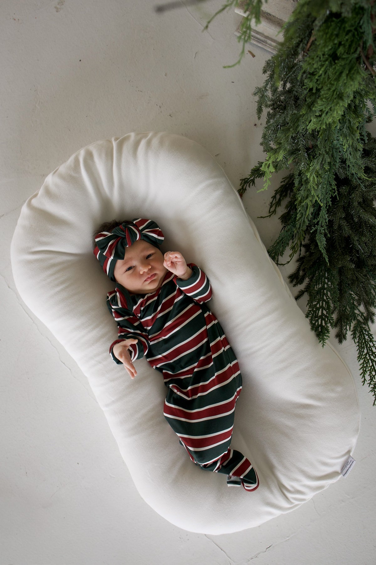 A newborn wearing a Bamboo Knotted Gown | Holiday Stripe and matching headband, lying in a cushioned baby lounger.
