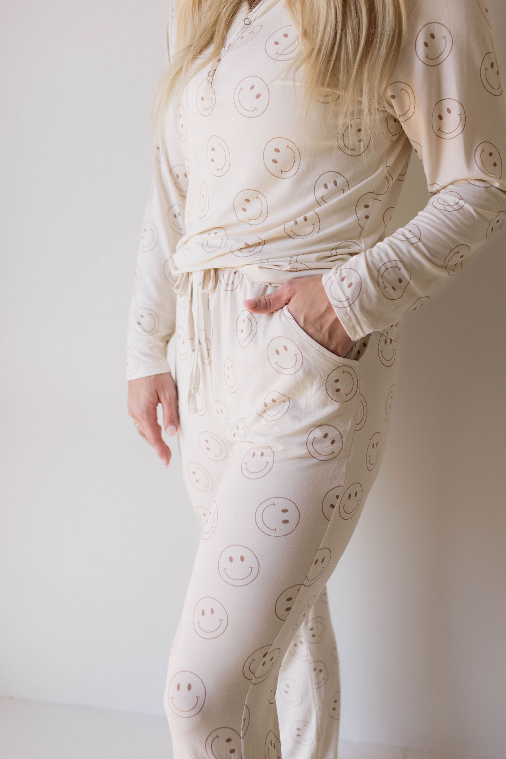 A person is standing against a plain background, wearing the Women's Bamboo Pajamas | Just Smile from forever french baby. The light-colored, long-sleeved pajamas set is adorned with a pattern of smiley faces and crafted from bamboo viscose, making it an ideal choice for breastfeeding moms. With one hand in their pocket and the other arm relaxed by their side, only the individual's torso, arms, and thighs are visible.