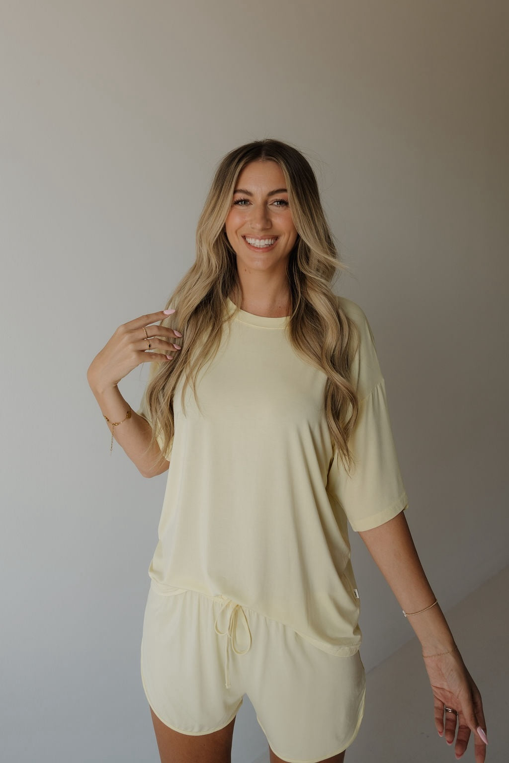 A woman with long blonde hair wears the forever french baby Women’s Bamboo Short Set in Sunny, featuring a loose, light yellow short-sleeve top and matching shorts made from breathable bamboo fabric. She smiles while standing against a plain, light background.